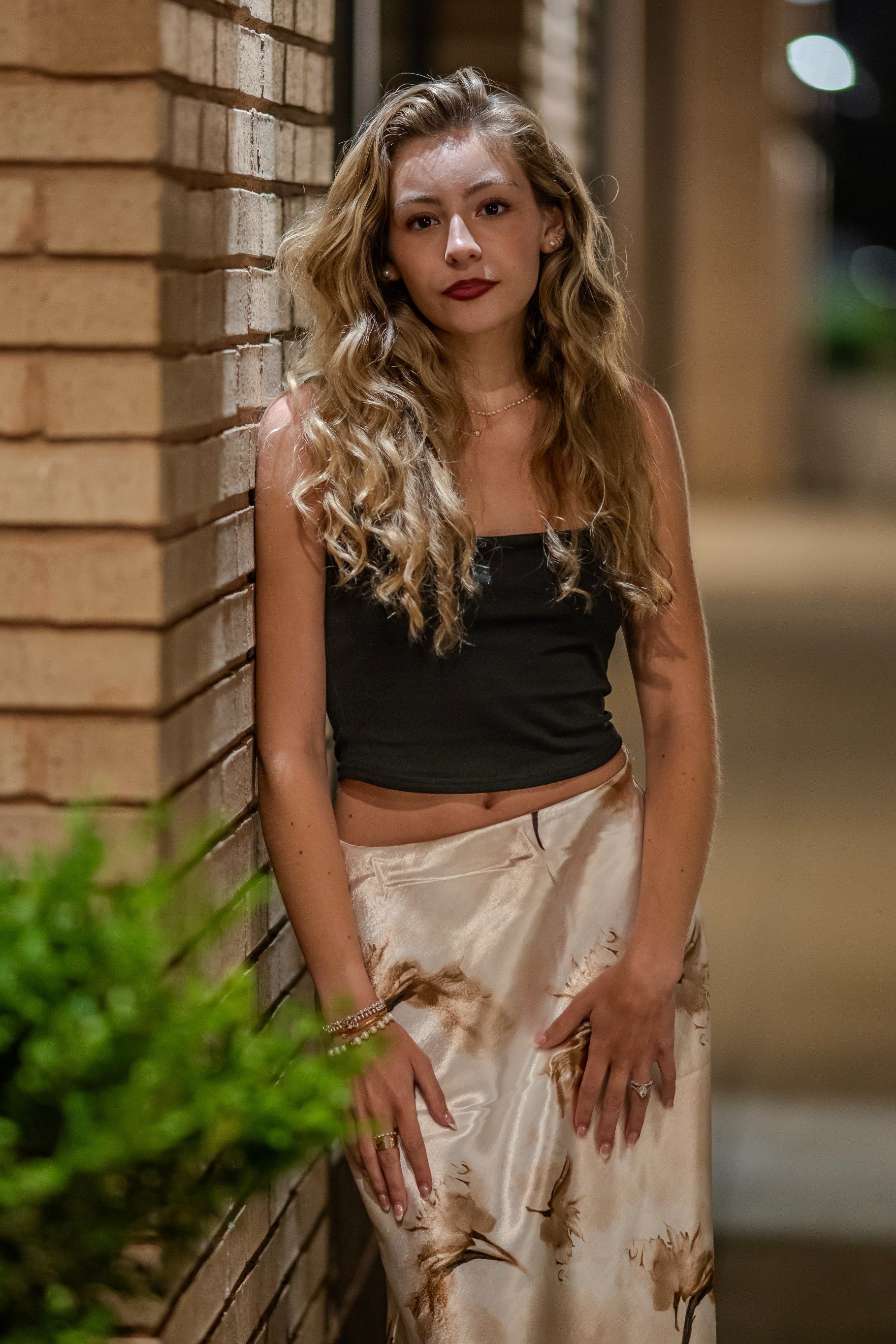 Woman standing by a brick wall at night, wearing a black top and beige satin skirt.