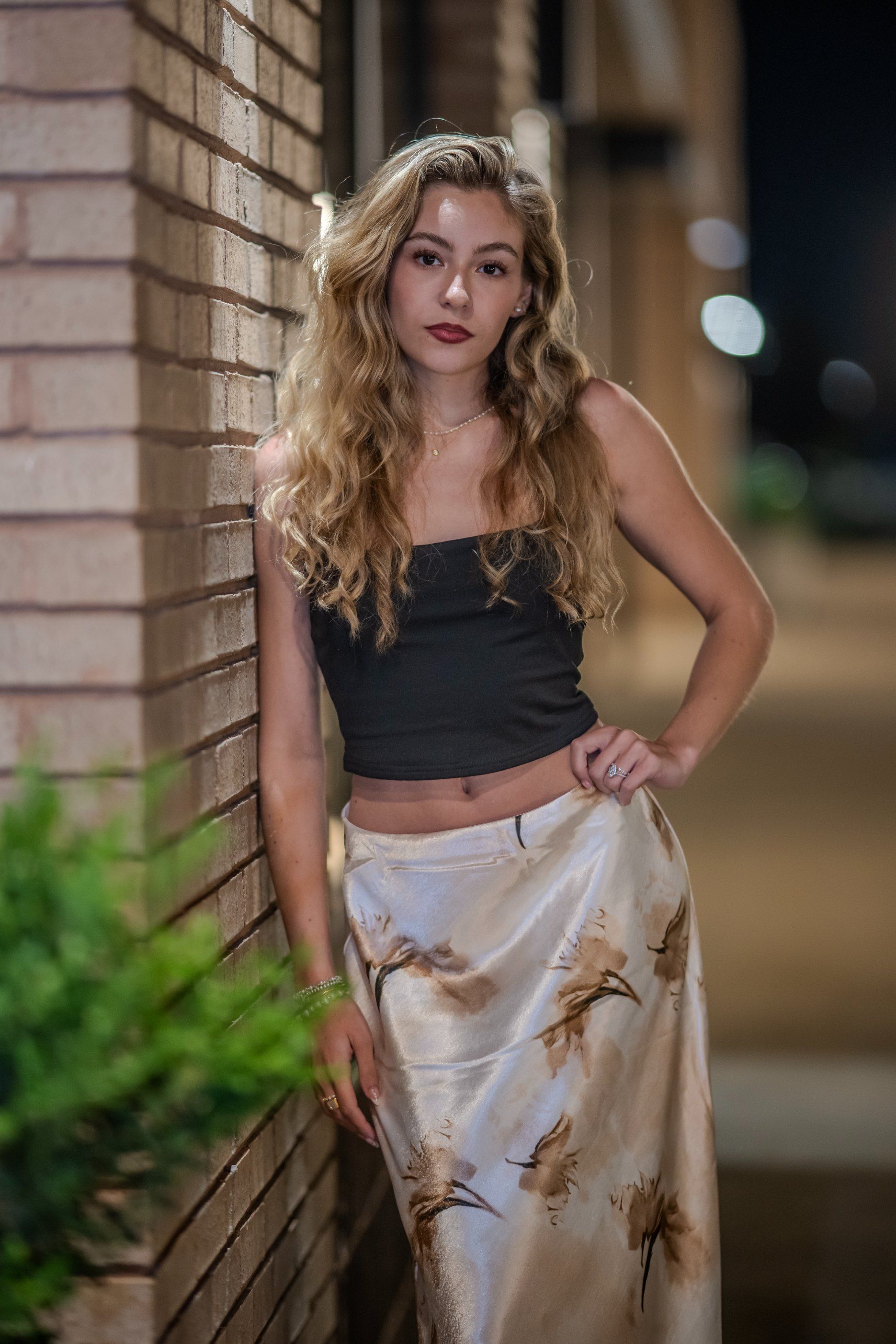 Woman leaning against a brick wall at night, wearing a black top and white floral skirt.