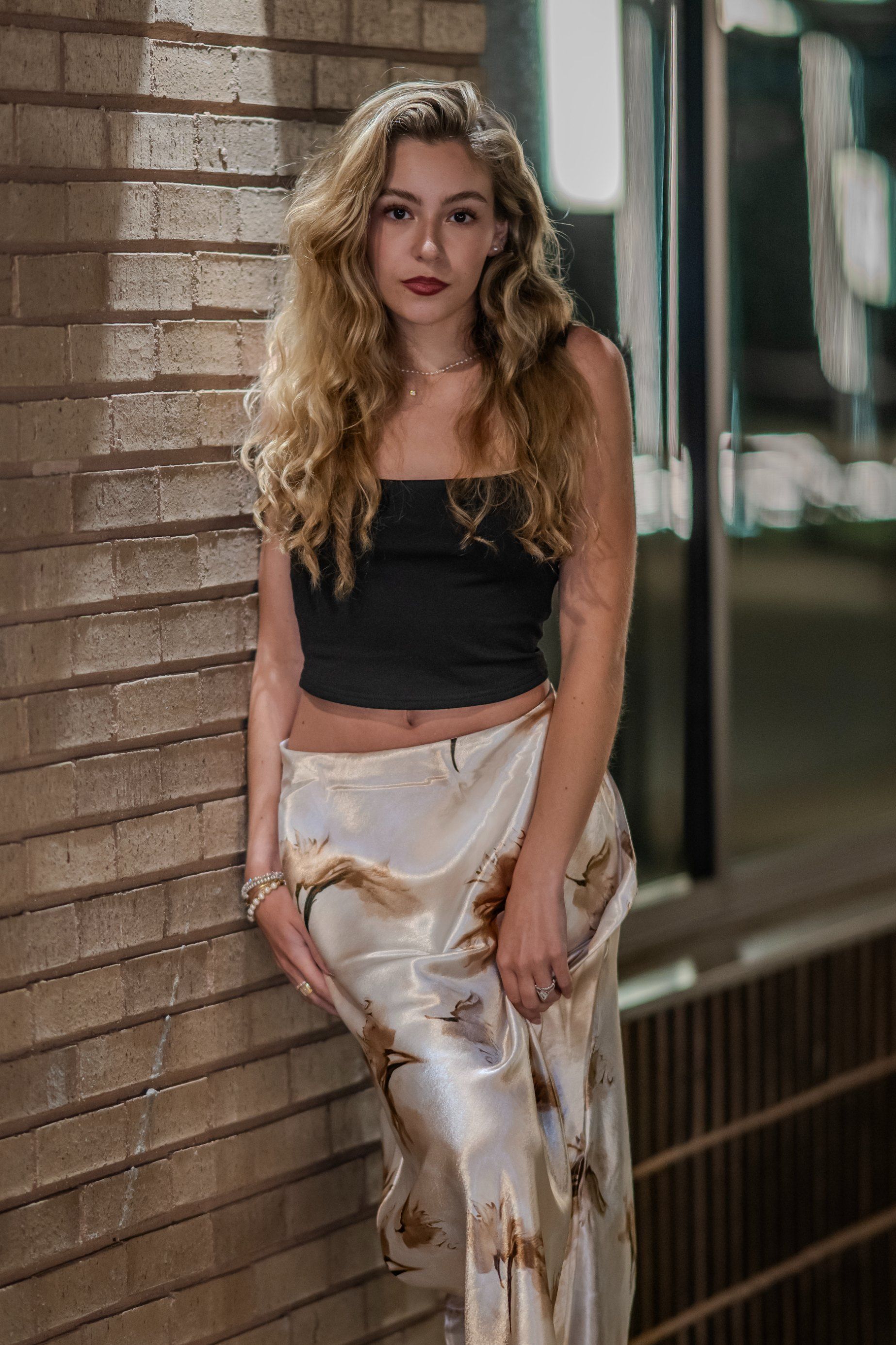 Woman in black top and white patterned pants standing by a brick wall indoors.
