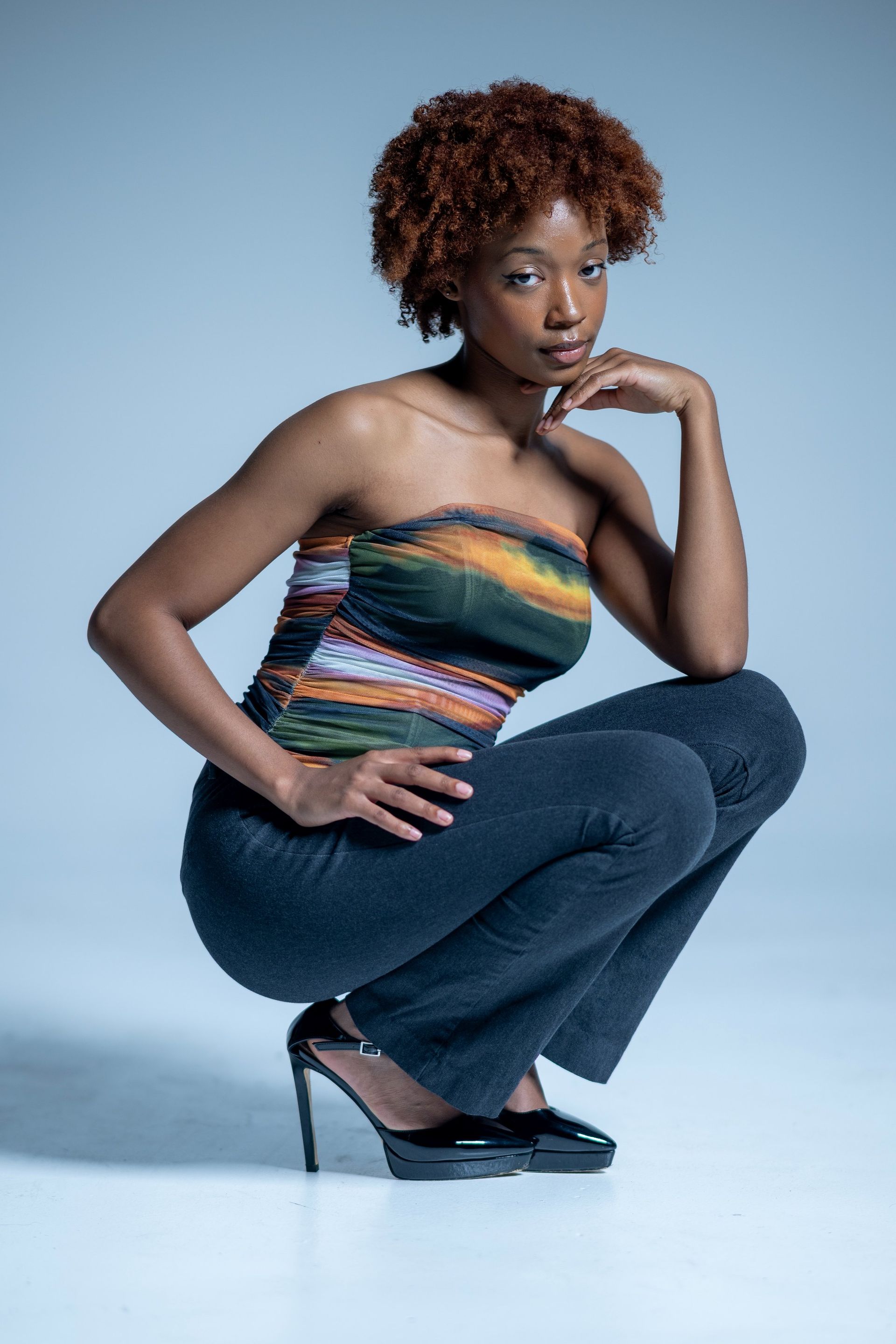 Woman crouching in colorful strapless top, dark jeans, and heels against a blue studio backdrop