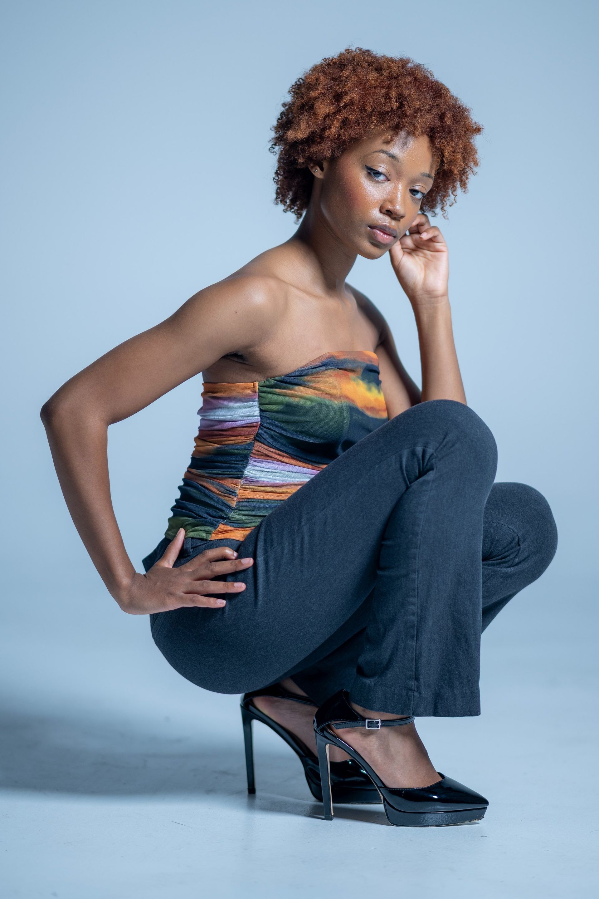 Woman crouching in colorful strapless top, dark jeans, and heels against a light blue studio background