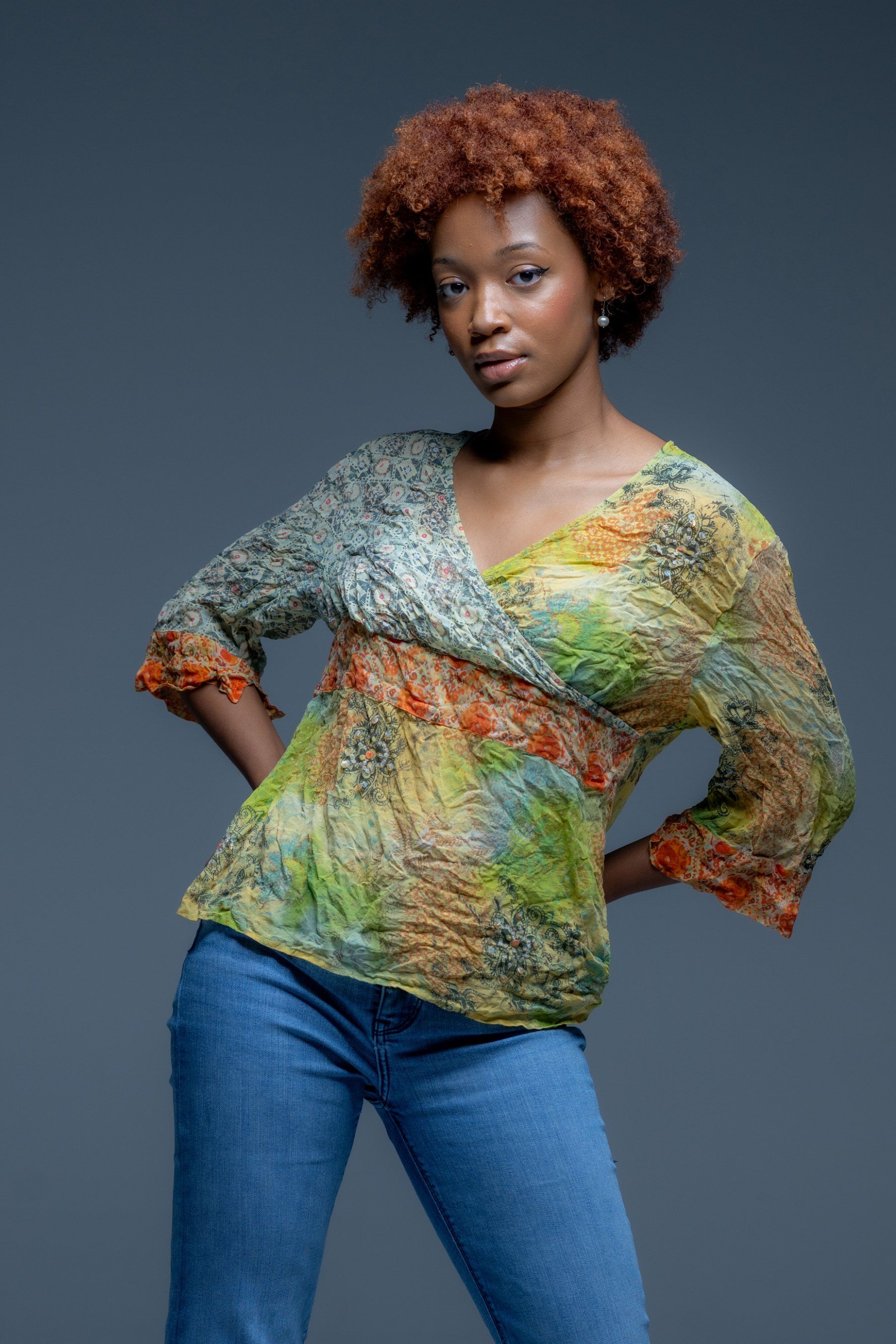 Person posing in a multicolored sheer top and jeans against a dark studio backdrop