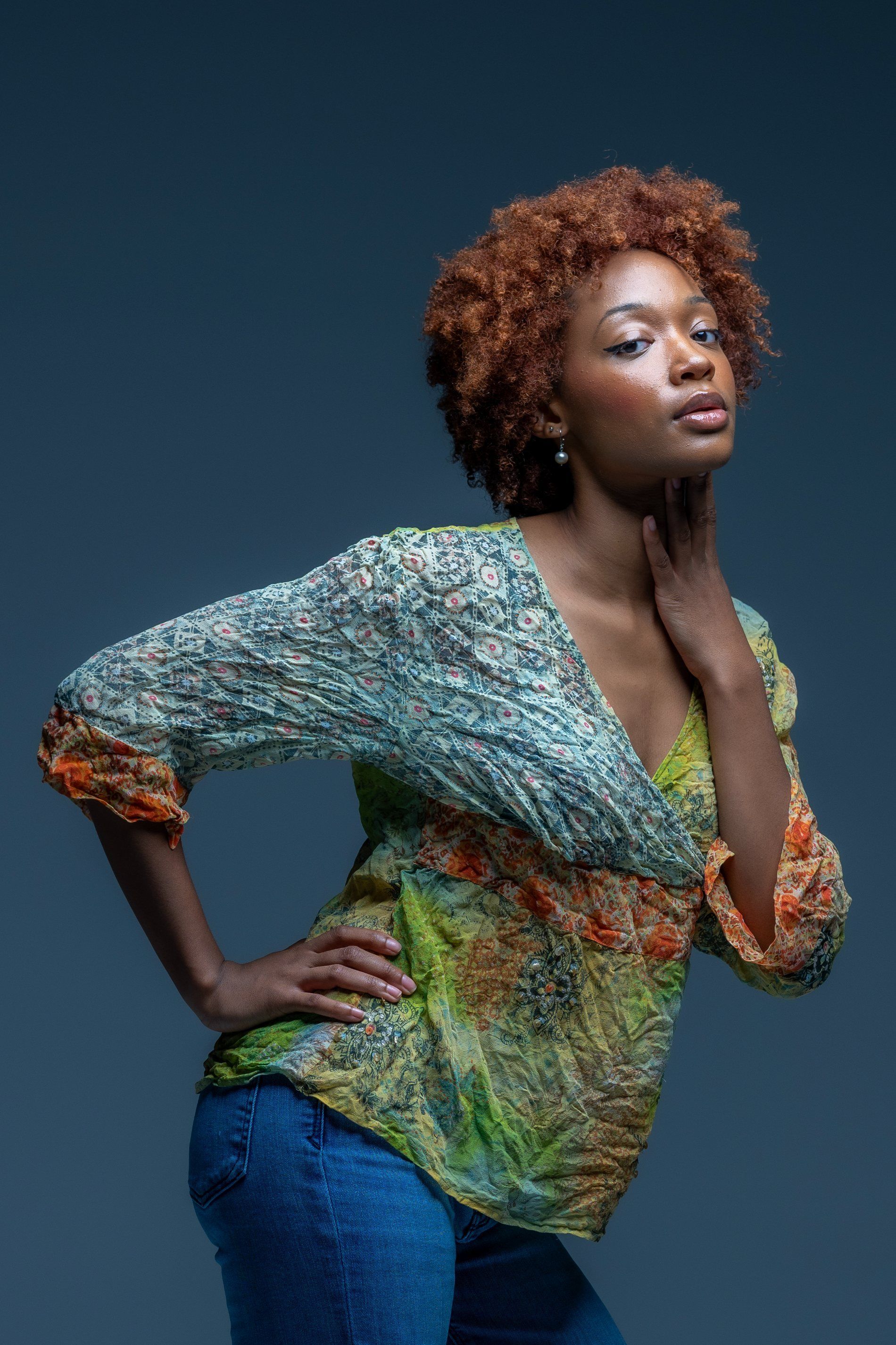 Portrait of a person posing against a dark blue background, wearing a colorful patterned top and blue jeans.
