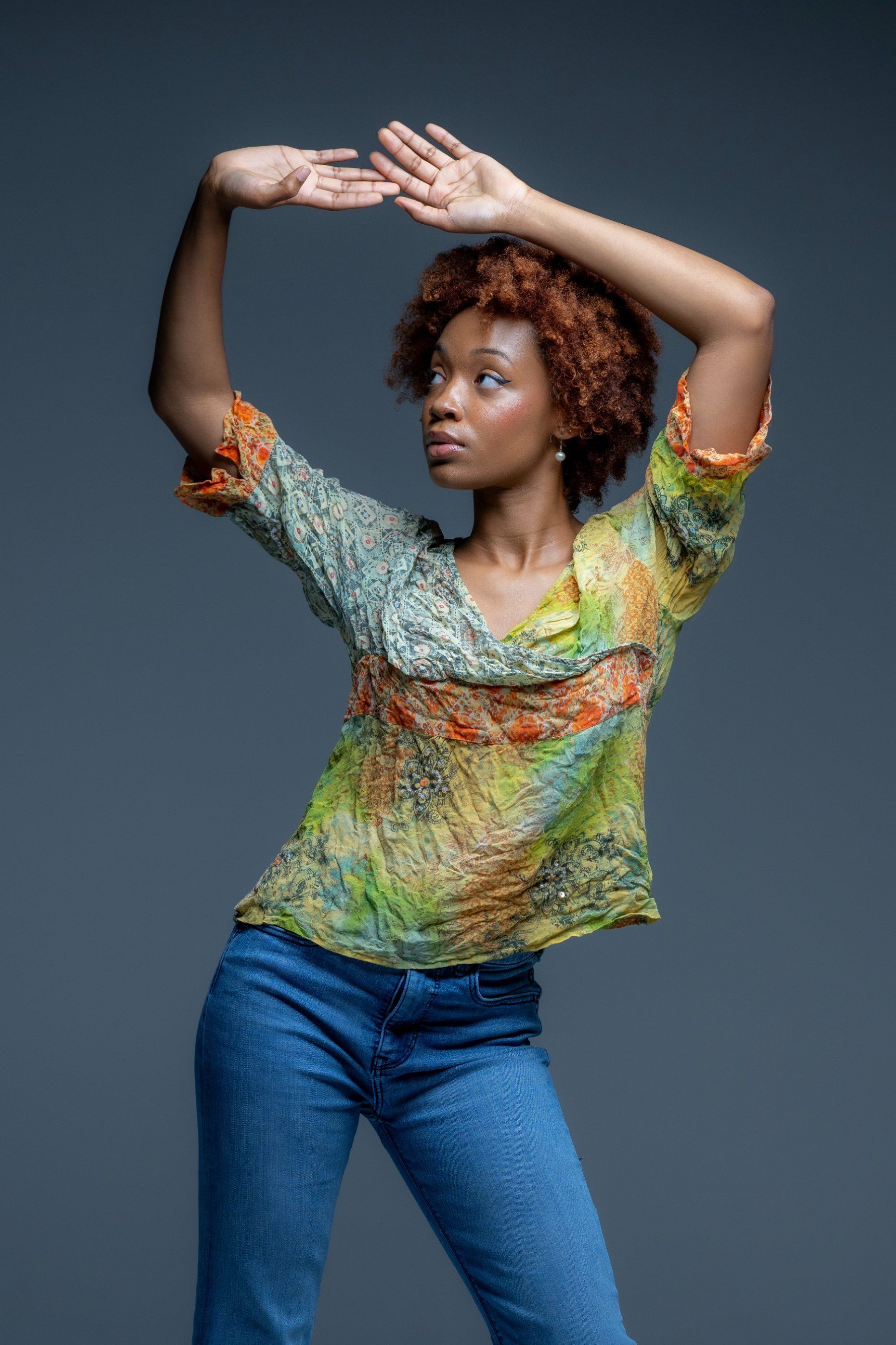 Person posing with arms raised, wearing a colorful top and blue jeans against a gray background