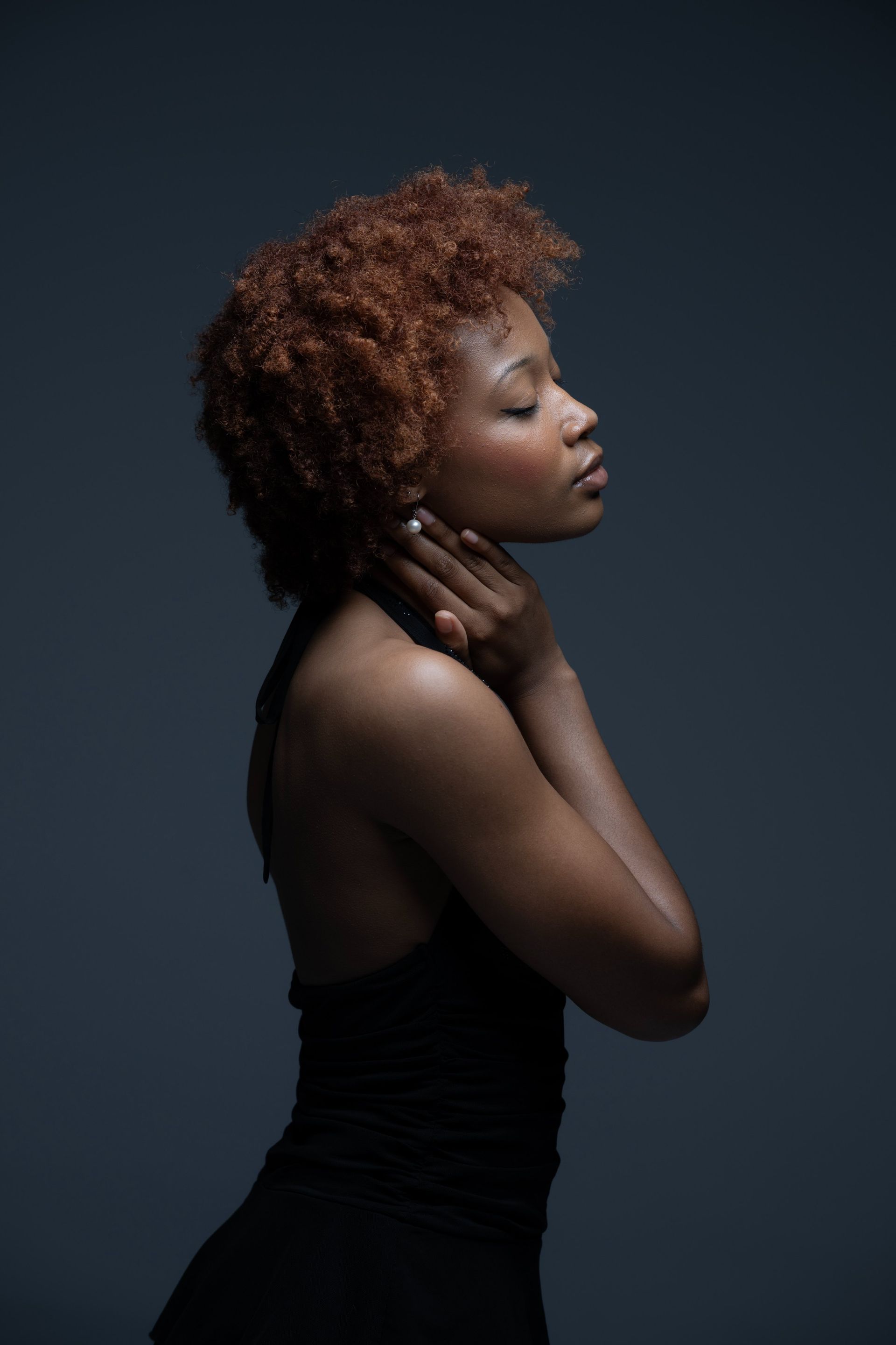 Side-profile portrait of a person in a black dress against a dark background, hands resting on neck.
