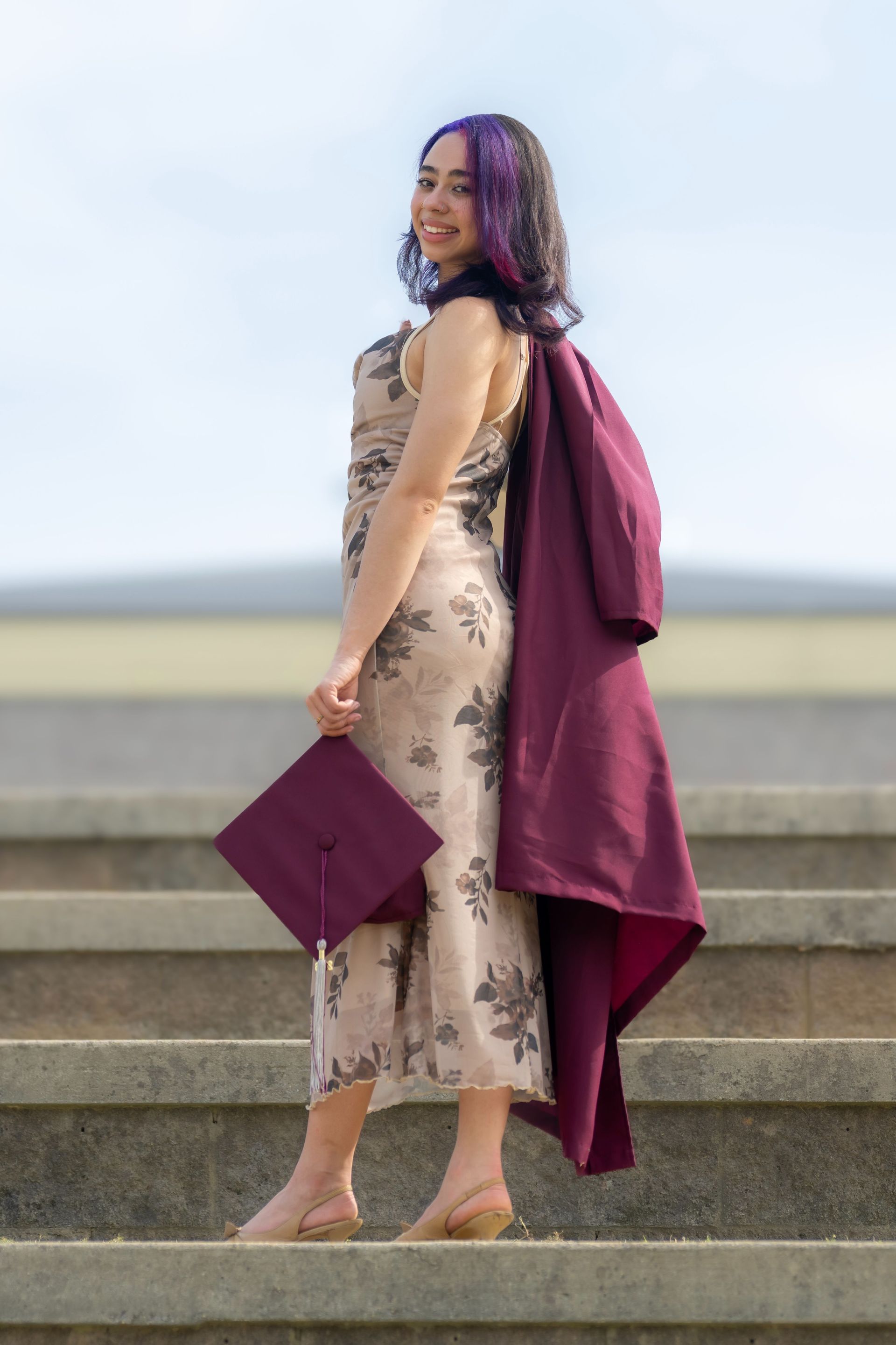 Woman in a beige floral dress with a burgundy shawl, standing on stone steps and holding a matching clutch.
