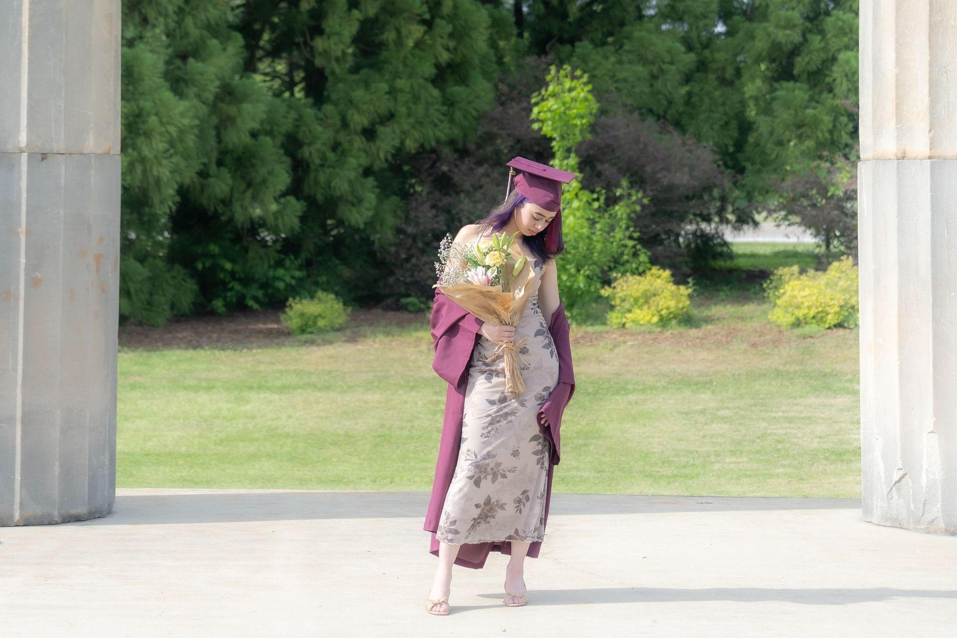 Graduate in maroon cap and gown poses outdoors between stone columns.