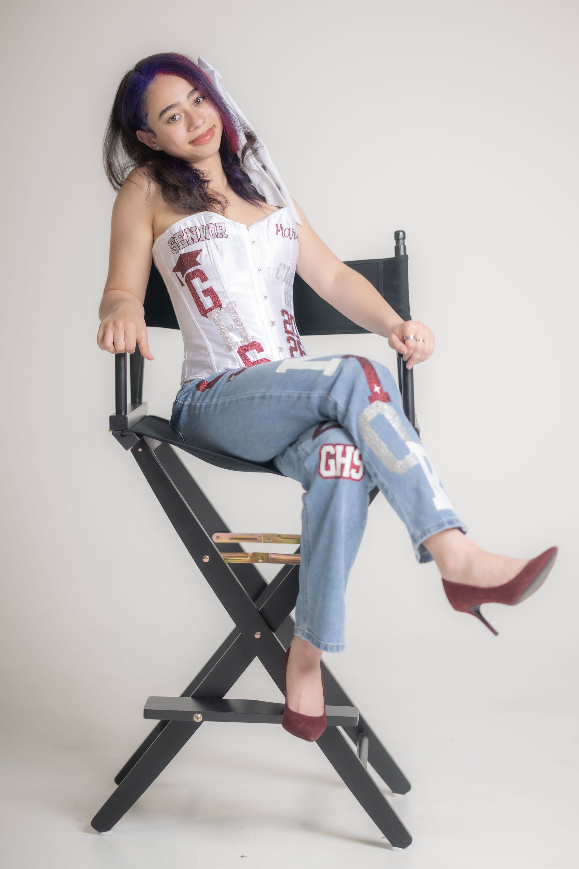 Woman seated on a director’s chair in a white studio, wearing a white top, jeans, and red heels.