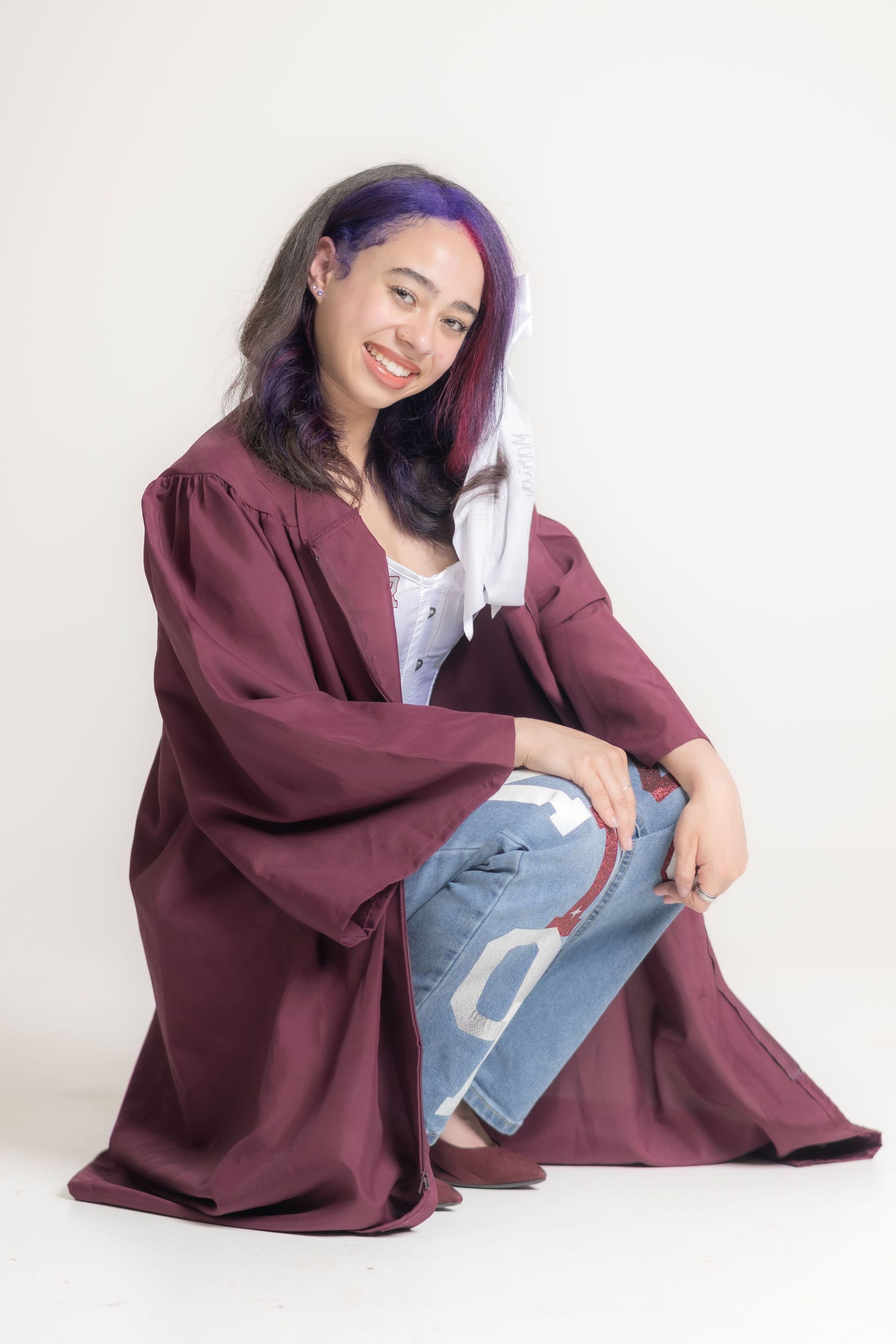 Person seated on a white background, wearing a maroon coat over a white top and ripped blue jeans.