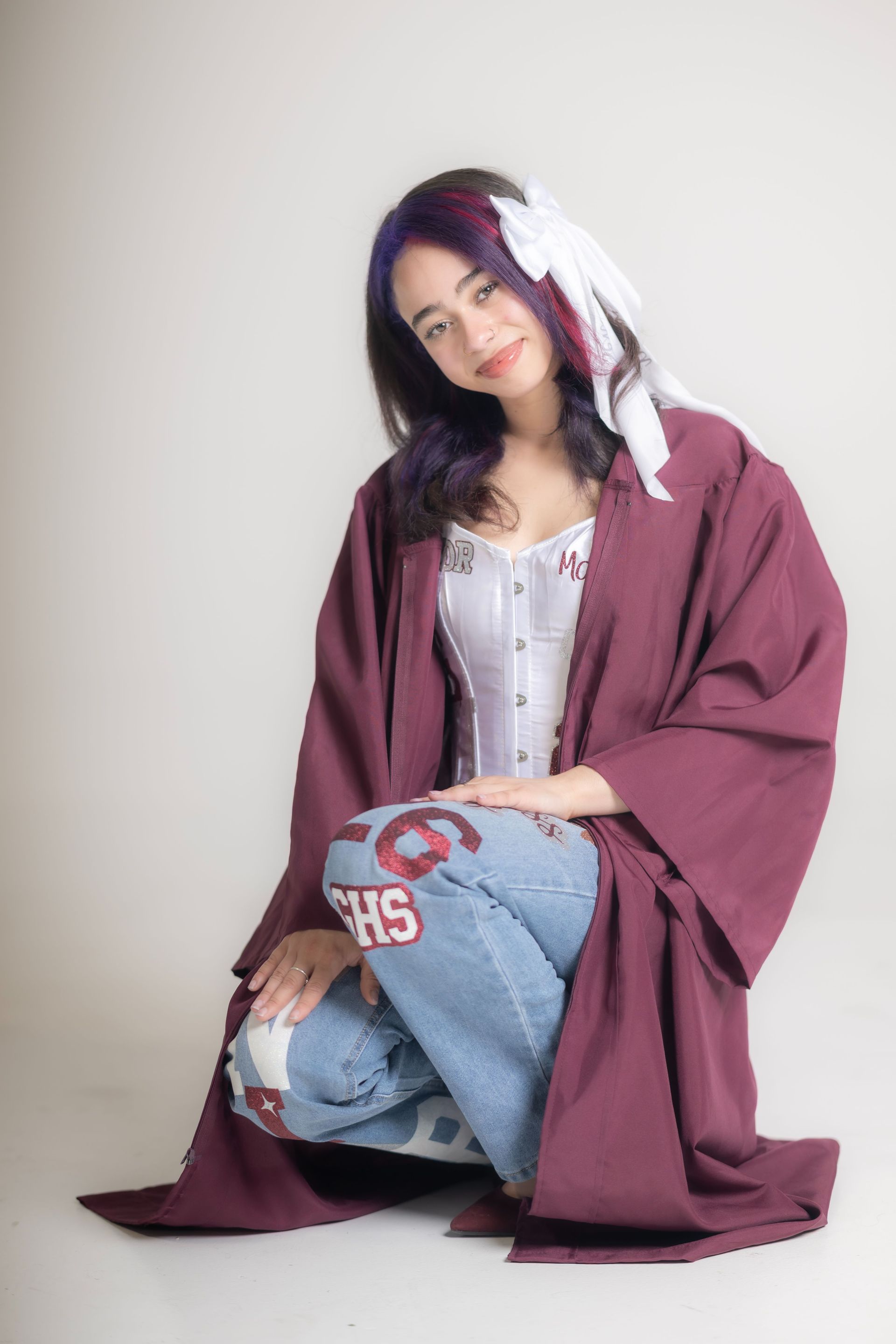 Person kneeling in a maroon coat and white top, smiling against a light backdrop, with a heart-covered pillow.