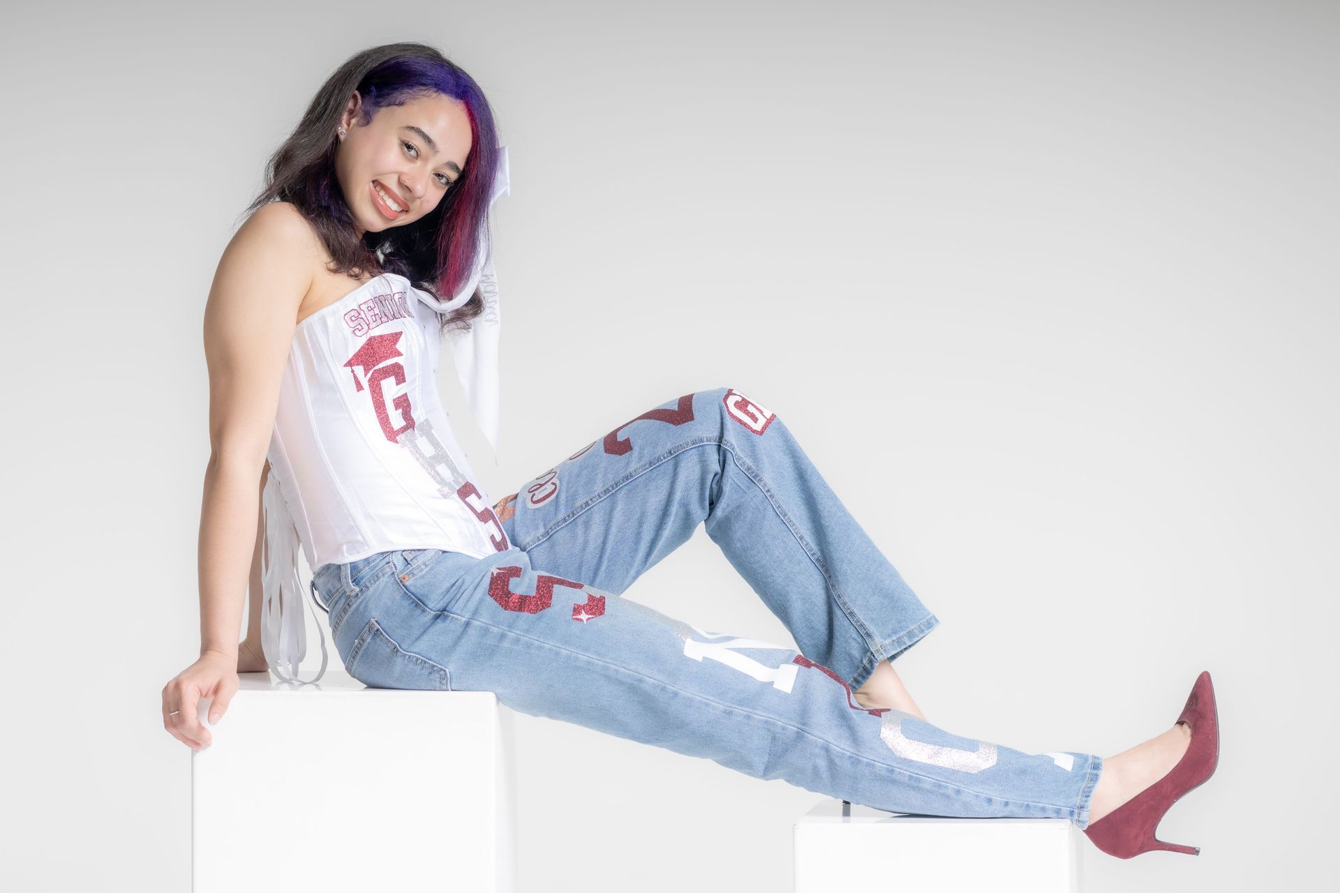 Person reclining on white blocks against a plain backdrop, wearing a white top, ripped jeans, and red heels.