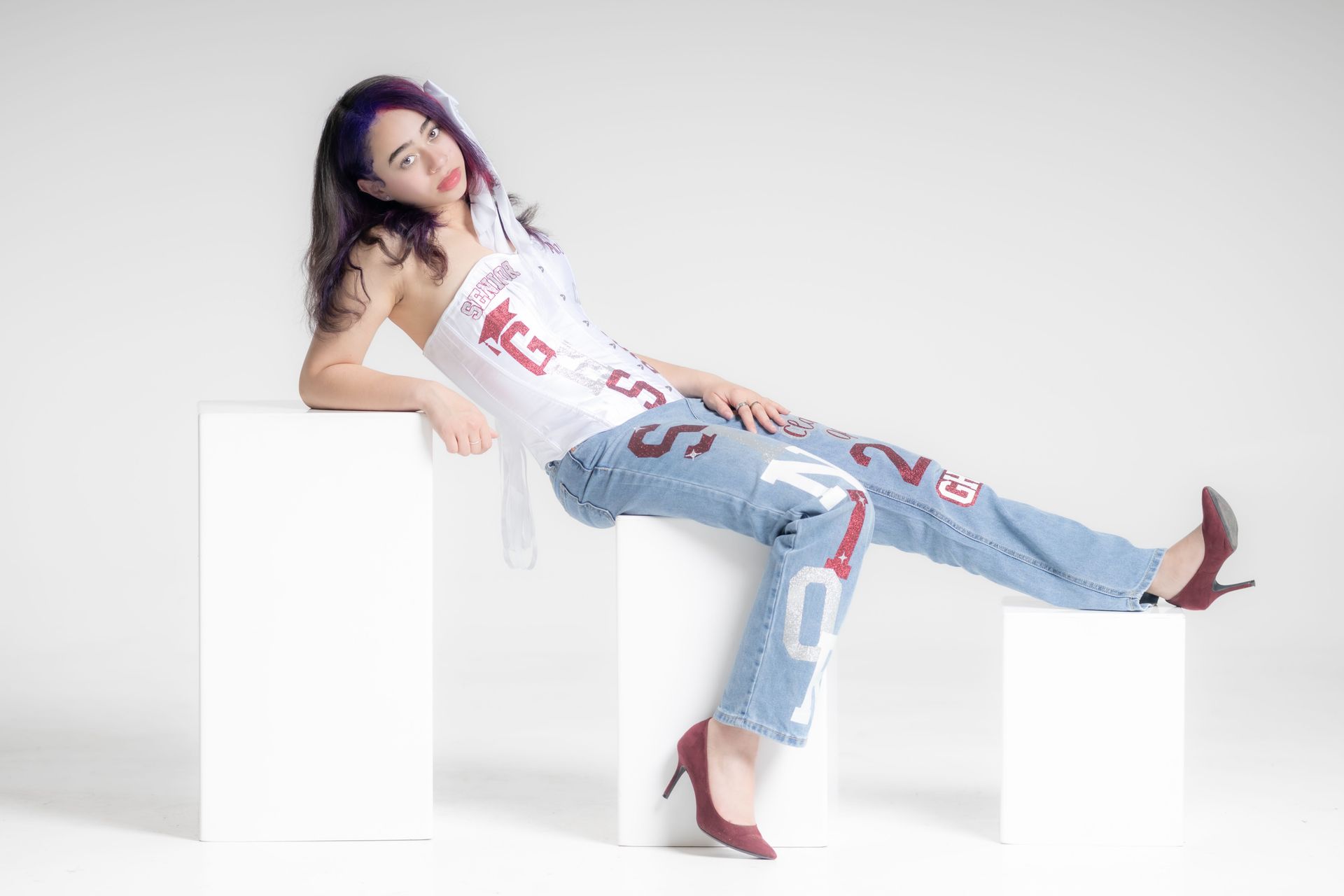 Woman reclining on white blocks, wearing a white graphic top and ripped jeans against a white background
