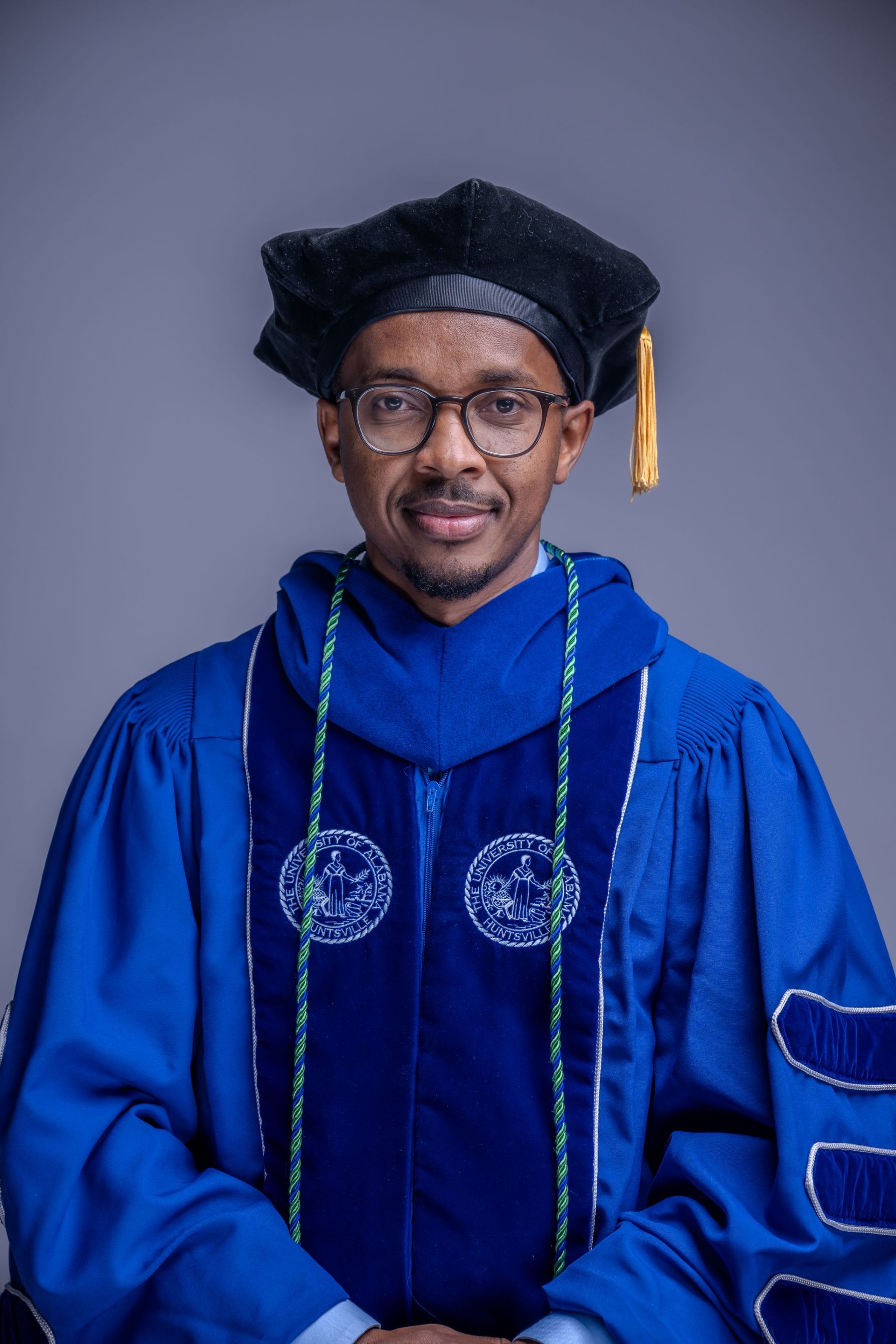 Portrait of a person in blue academic regalia and cap, smiling against a gray background