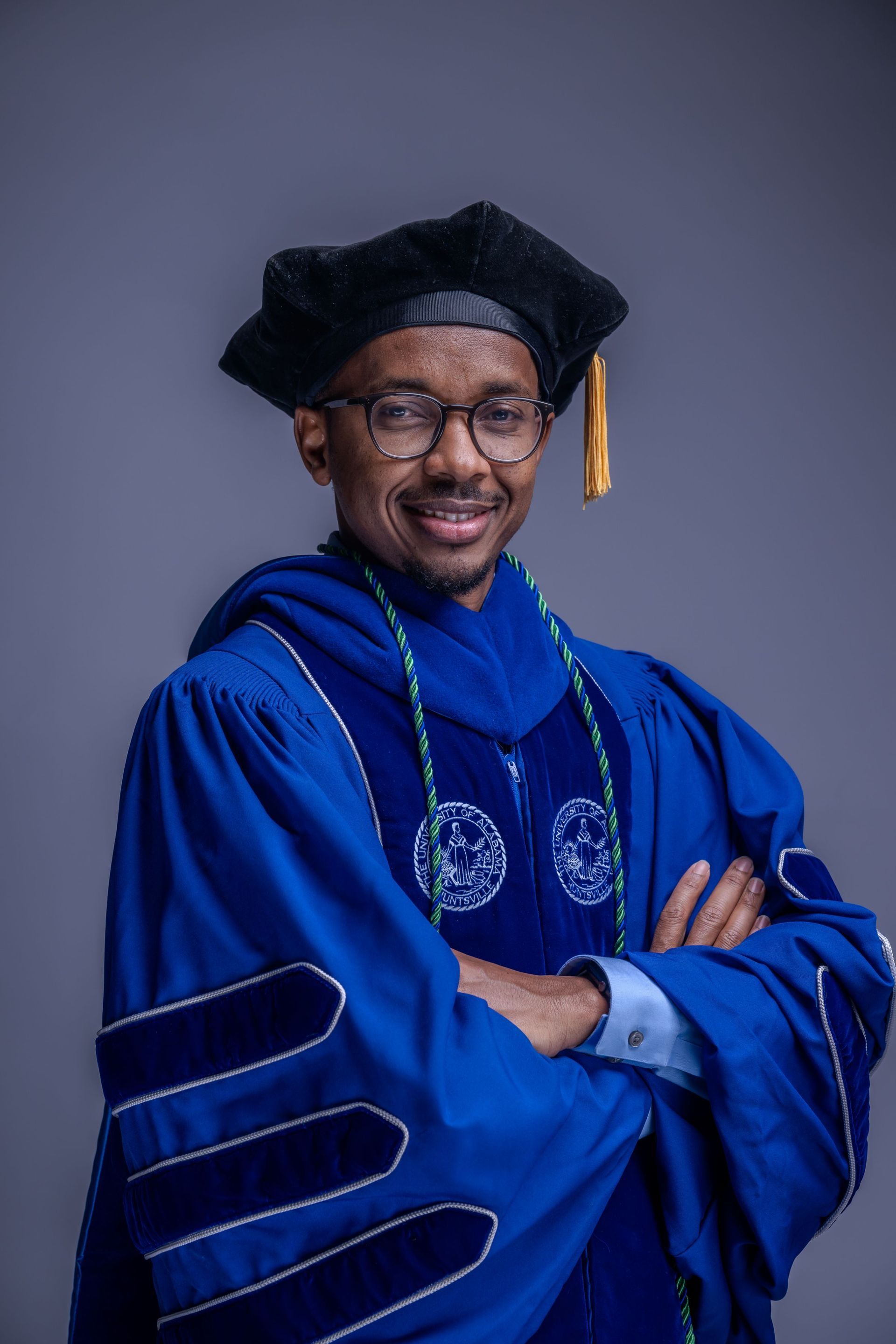 Person in blue graduation gown and cap, smiling with arms crossed against a gray background.