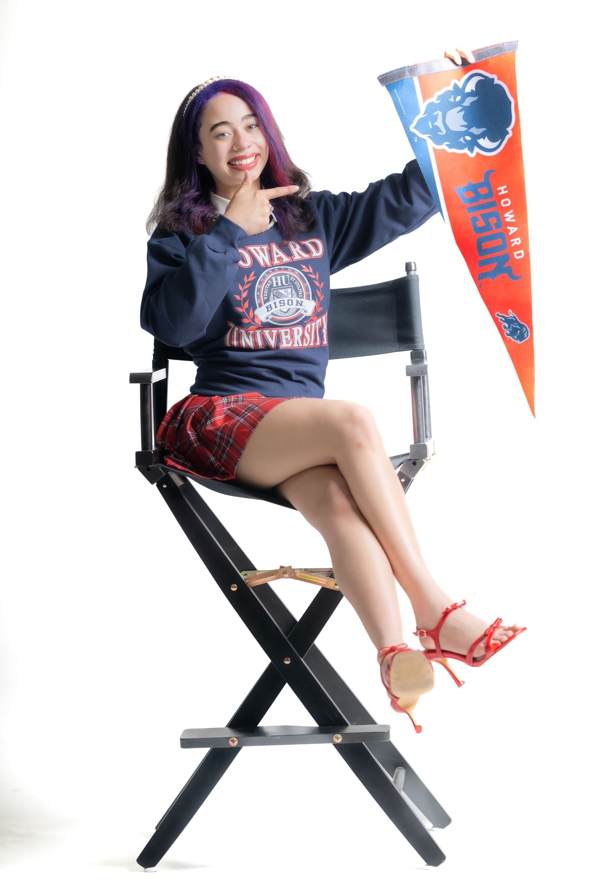 Woman seated on a director’s chair, holding an orange and blue pennant, wearing a navy sweater and red skirt.