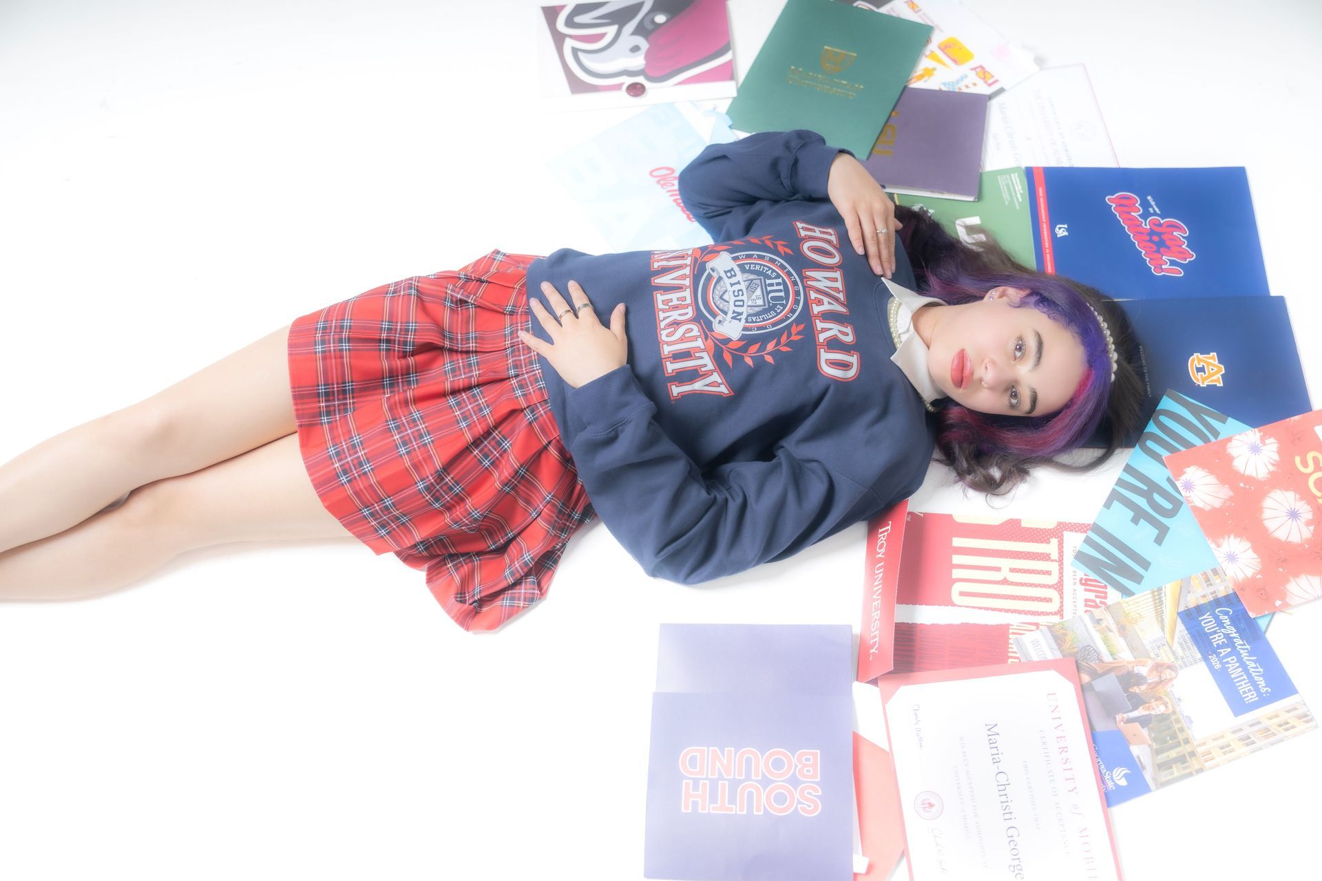 Person lying on the floor in a navy hoodie and red plaid skirt, surrounded by colorful books and notebooks.