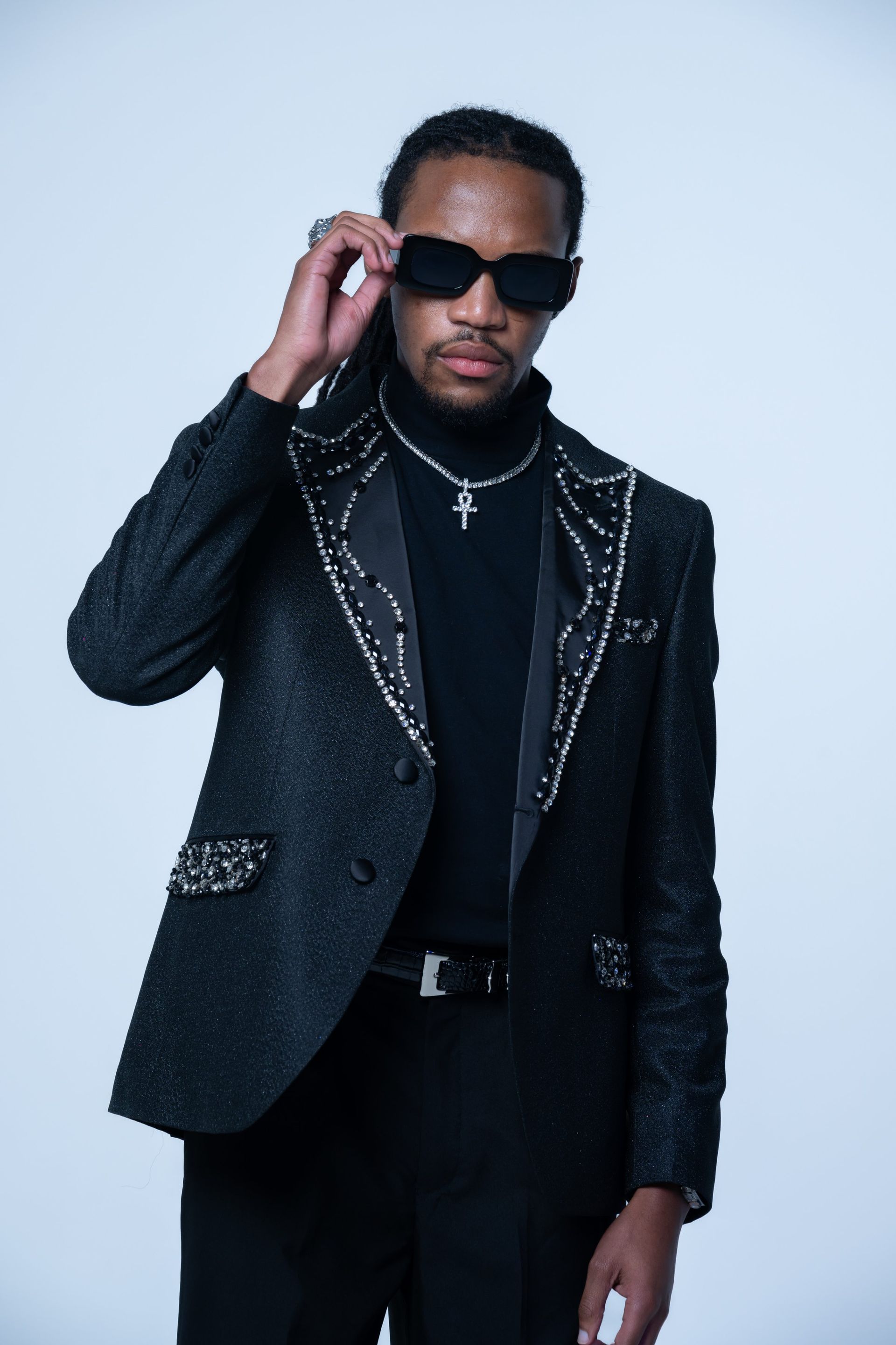 Man in black suit and turtleneck adjusting sunglasses against a light gray backdrop.