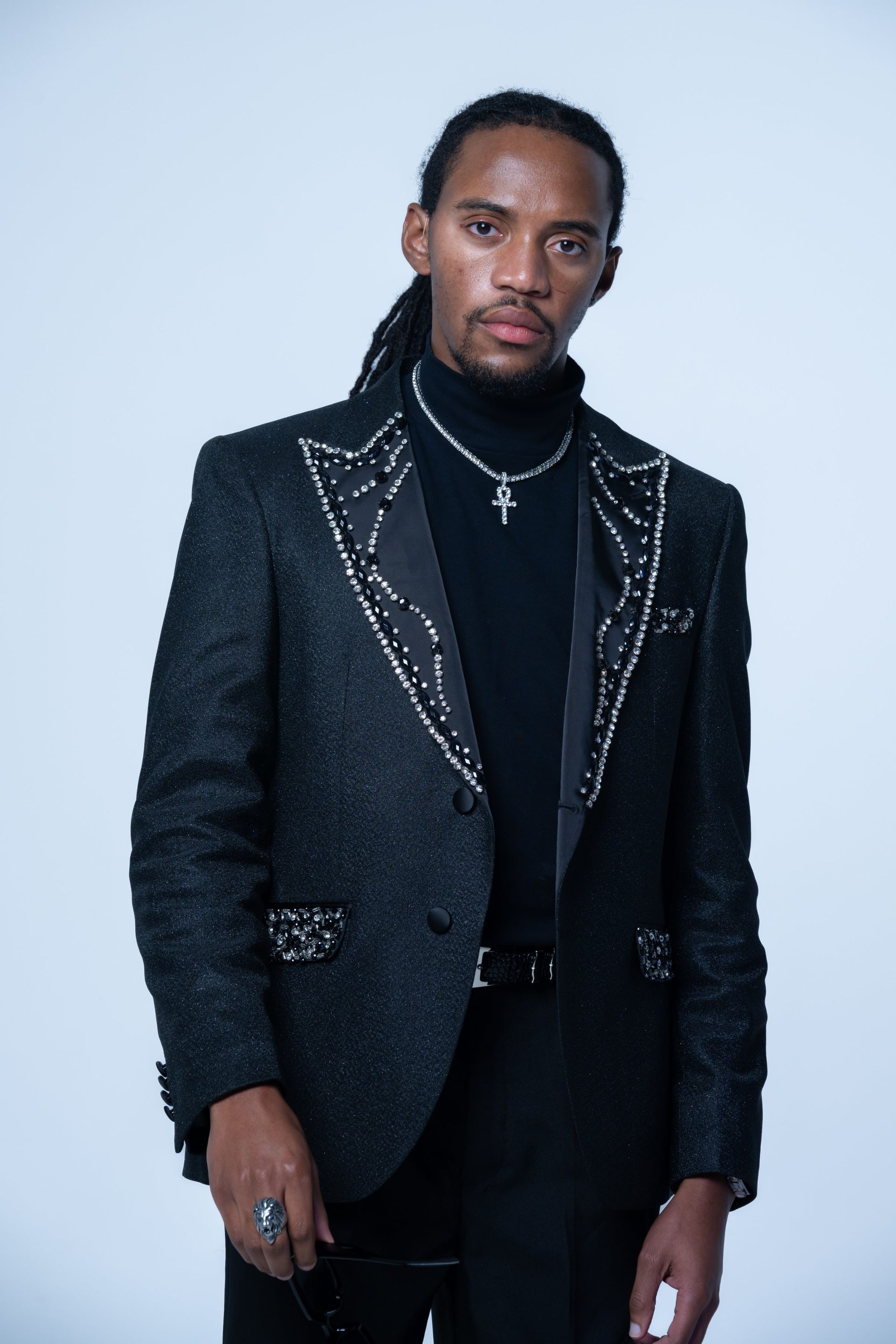 Man in a textured dark blazer over a black turtleneck, standing against a light blue background.