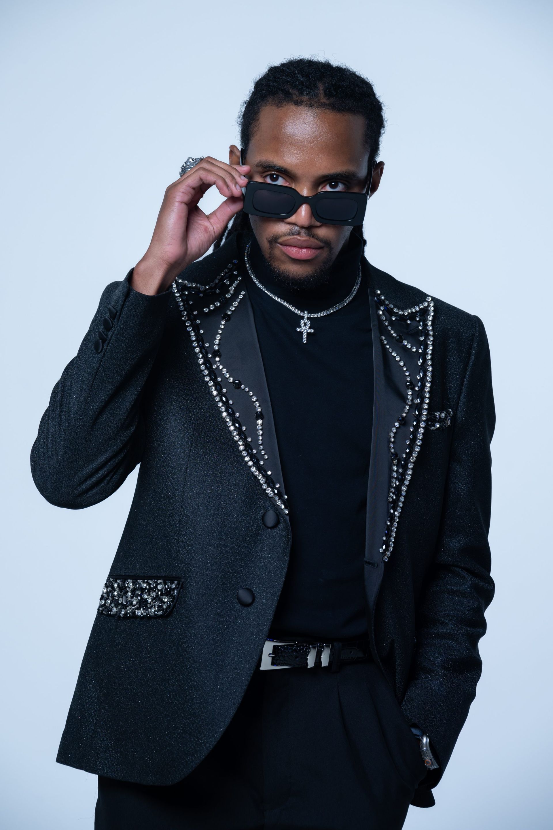 Man in a dark embellished suit adjusting sunglasses against a light background