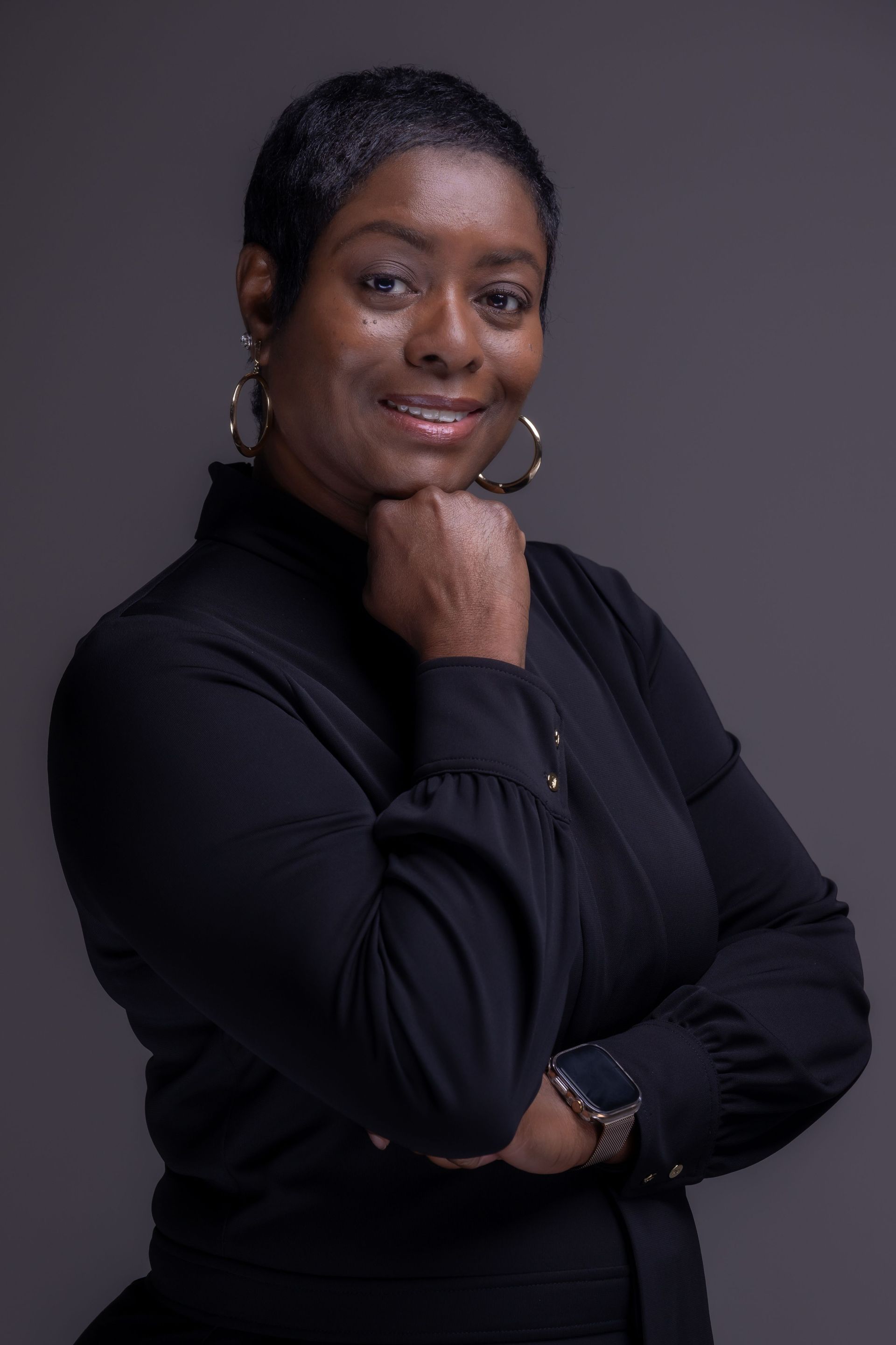 Portrait of a woman in a black top, smiling with her hand under her chin against a gray background