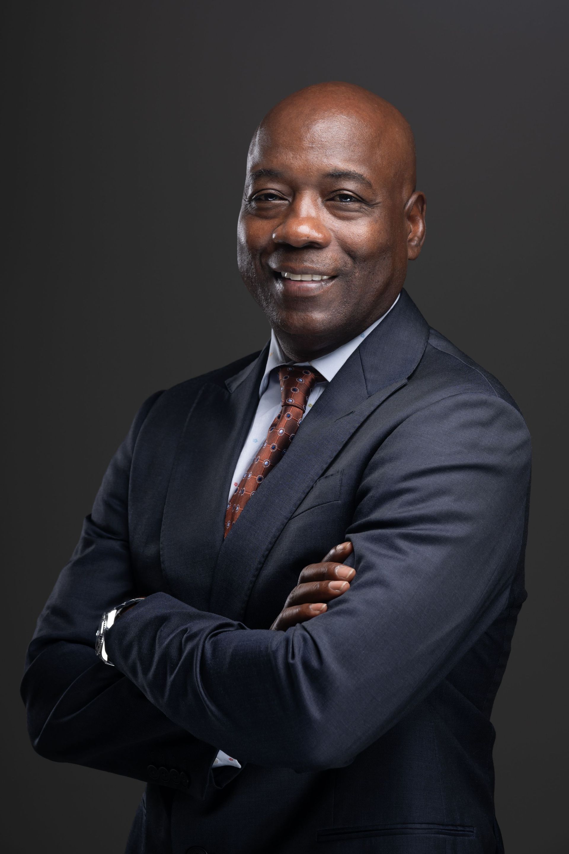 Smiling man in a dark suit with arms crossed against a gray studio backdrop