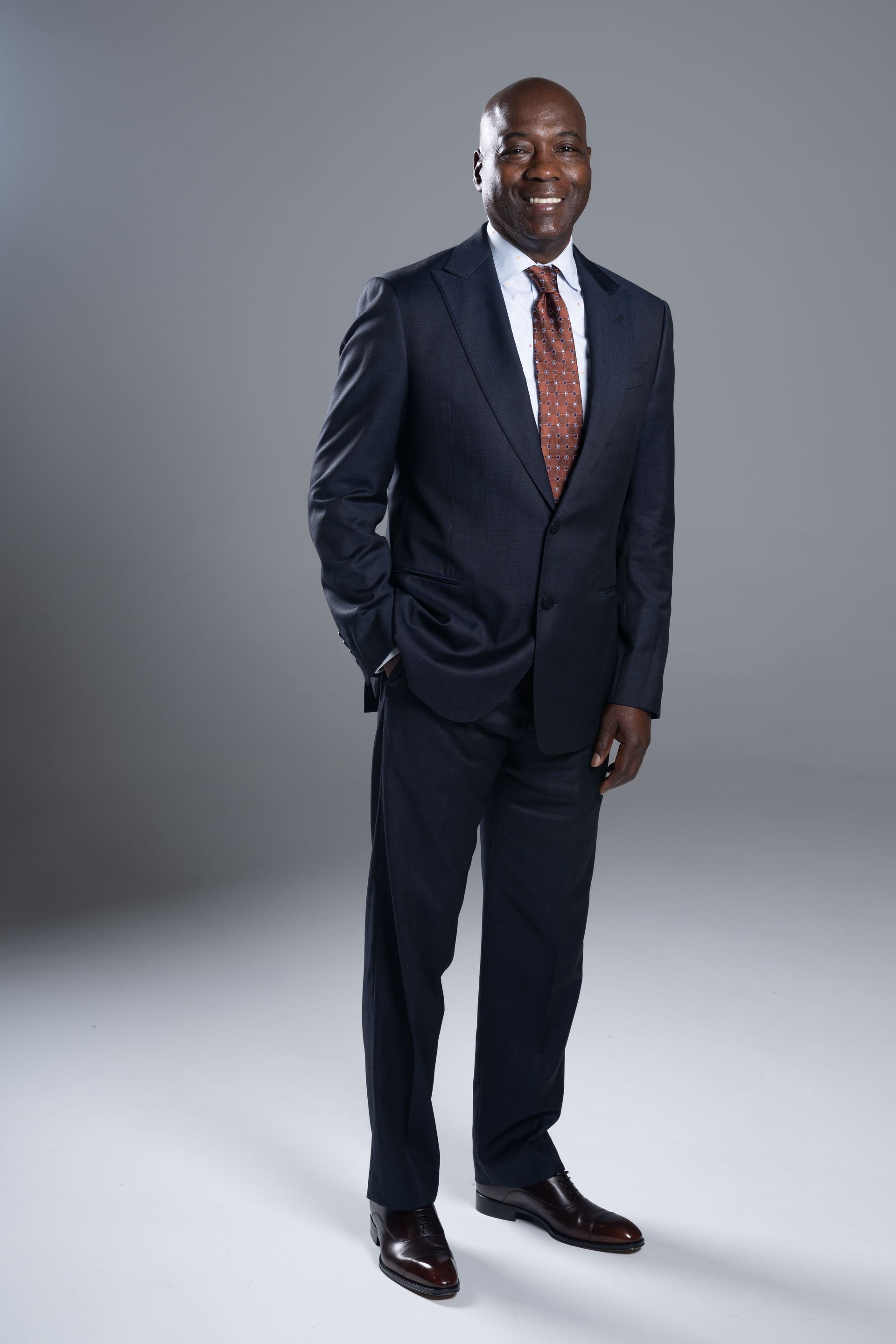 Man in a navy suit and red tie standing against a gray studio backdrop, smiling.
