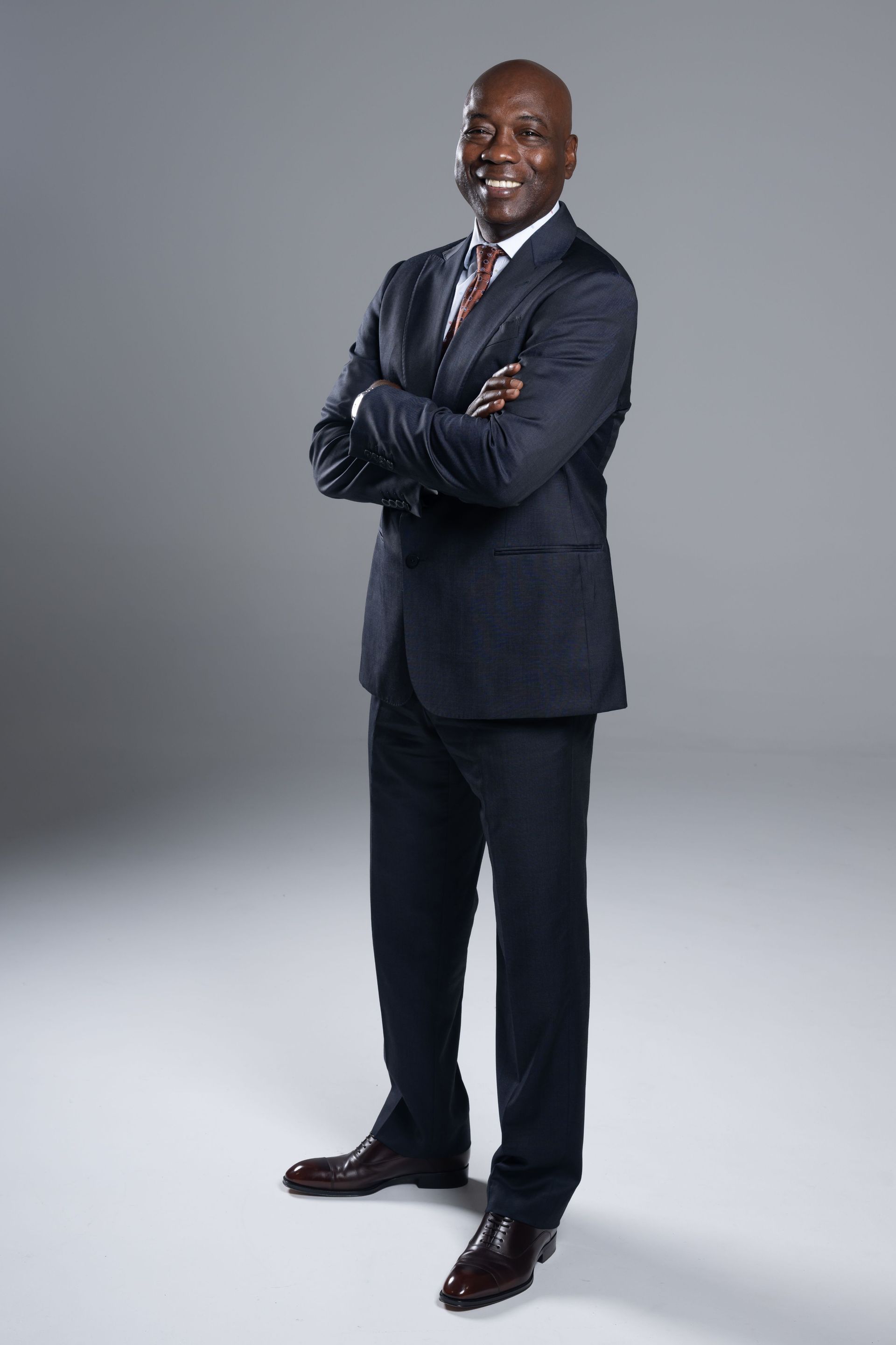 Full-length portrait of a man in a dark suit standing with arms crossed against a gray studio backdrop