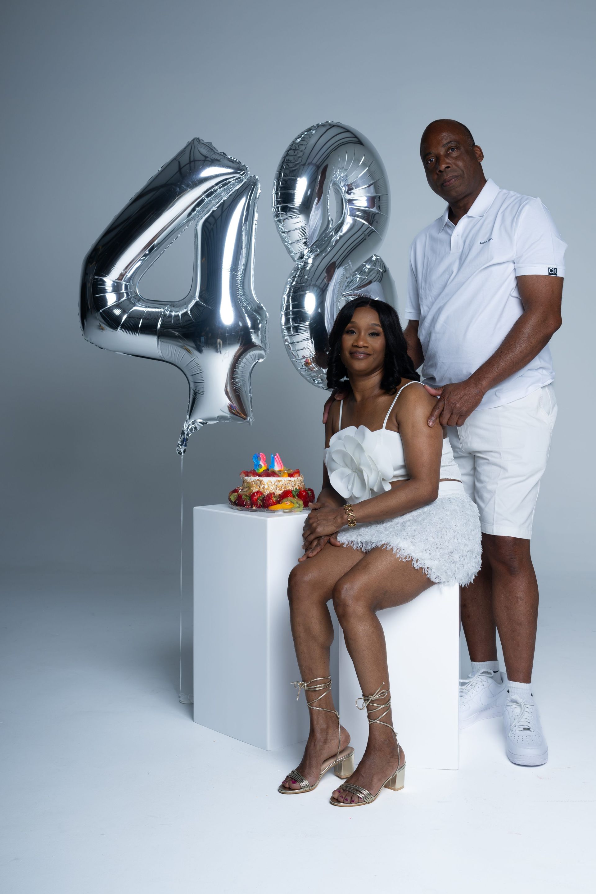 Couple posing beside silver “48” balloons and a birthday cake on a white pedestal