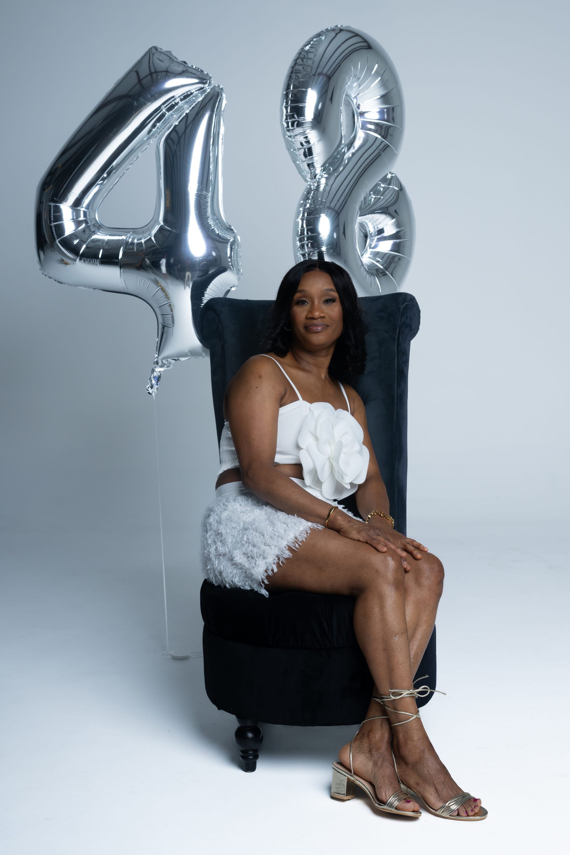 Woman seated on a black stool in a white dress with silver “48” balloons behind her.