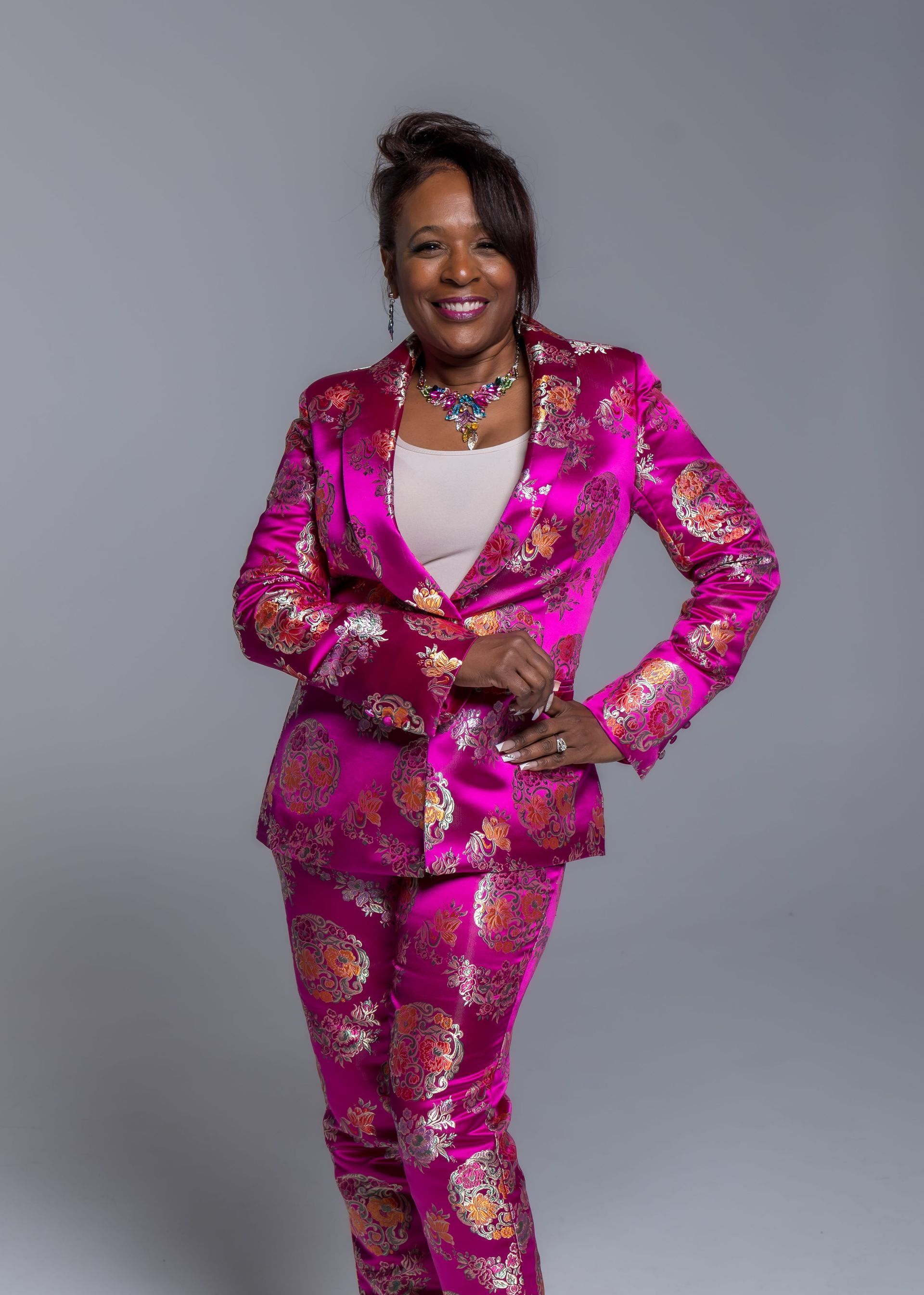 Person in a bright pink patterned suit poses against a gray studio backdrop.