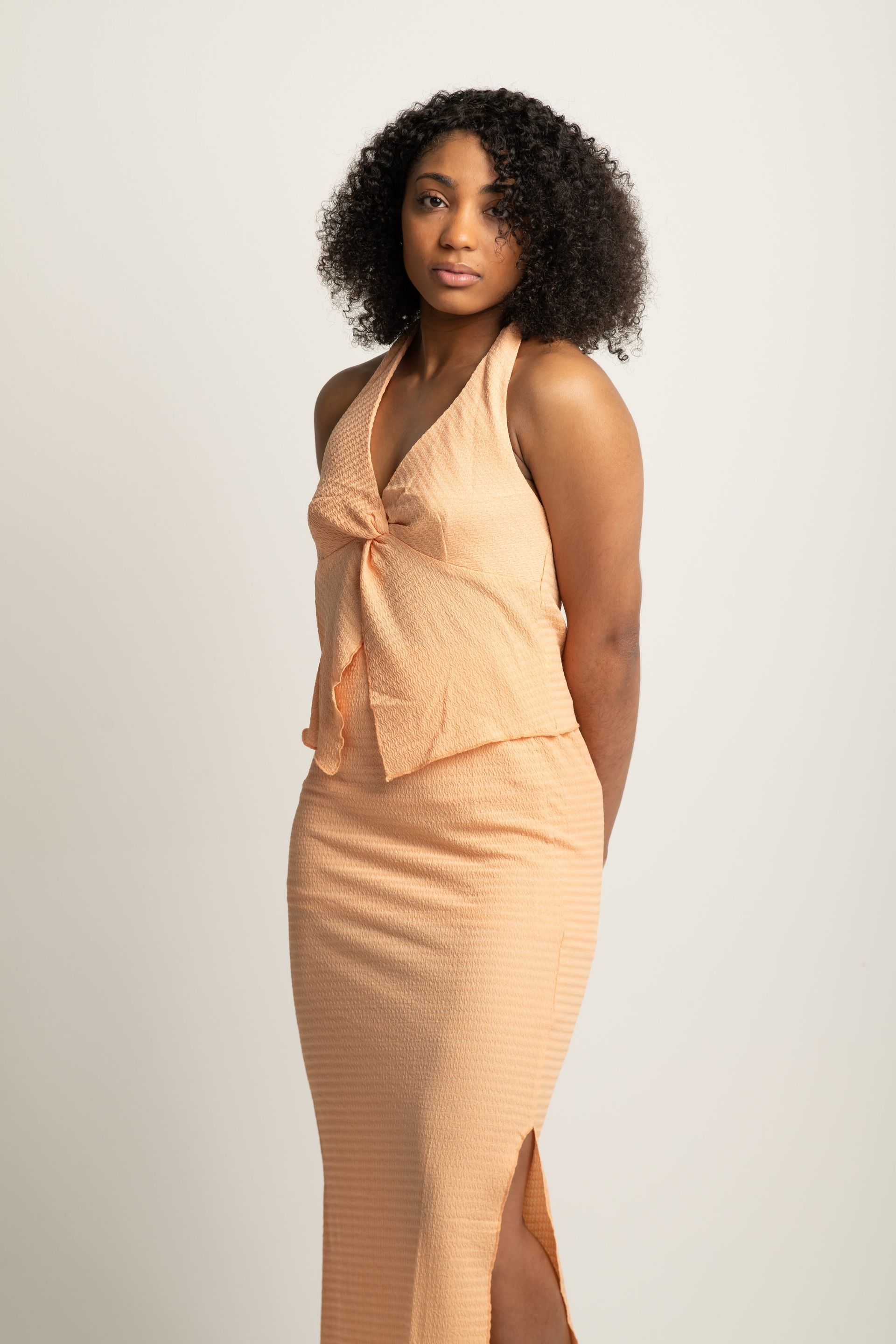 Woman in a peach dress posing against a light background, standing with hands behind her back.