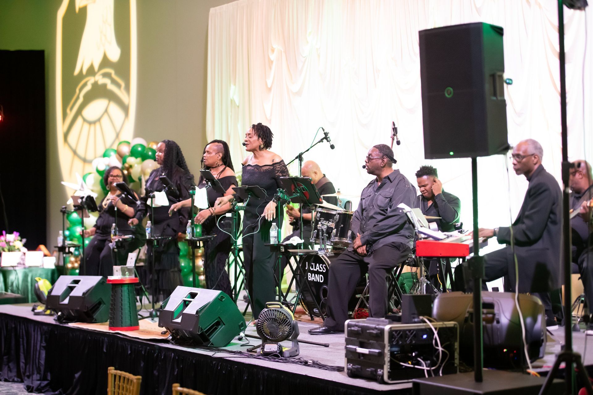 Band performing on a stage with microphones, instruments, and a large speaker in the background.