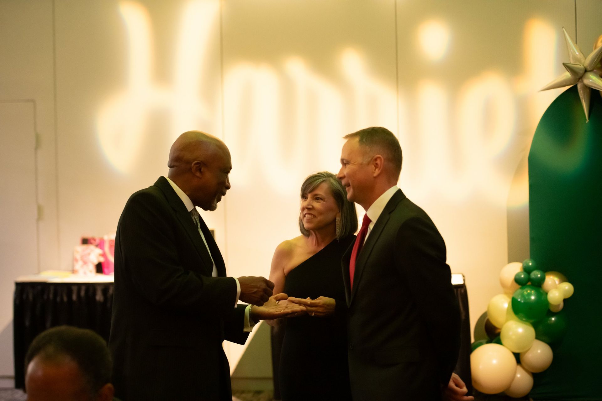 Three people in formal wear conversing at an indoor event with balloons and warm lighting.