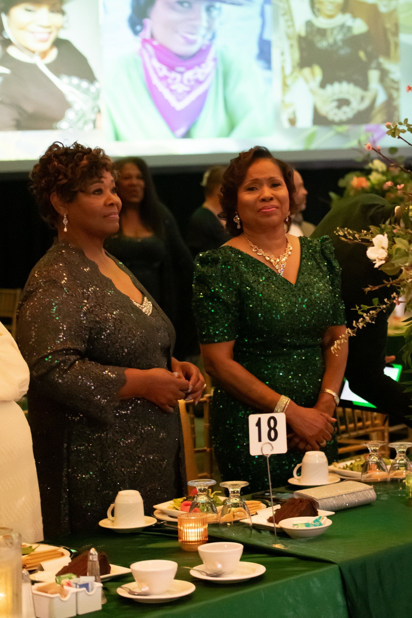 Two women in sparkly dresses standing at a decorated banquet table with a number 18 sign