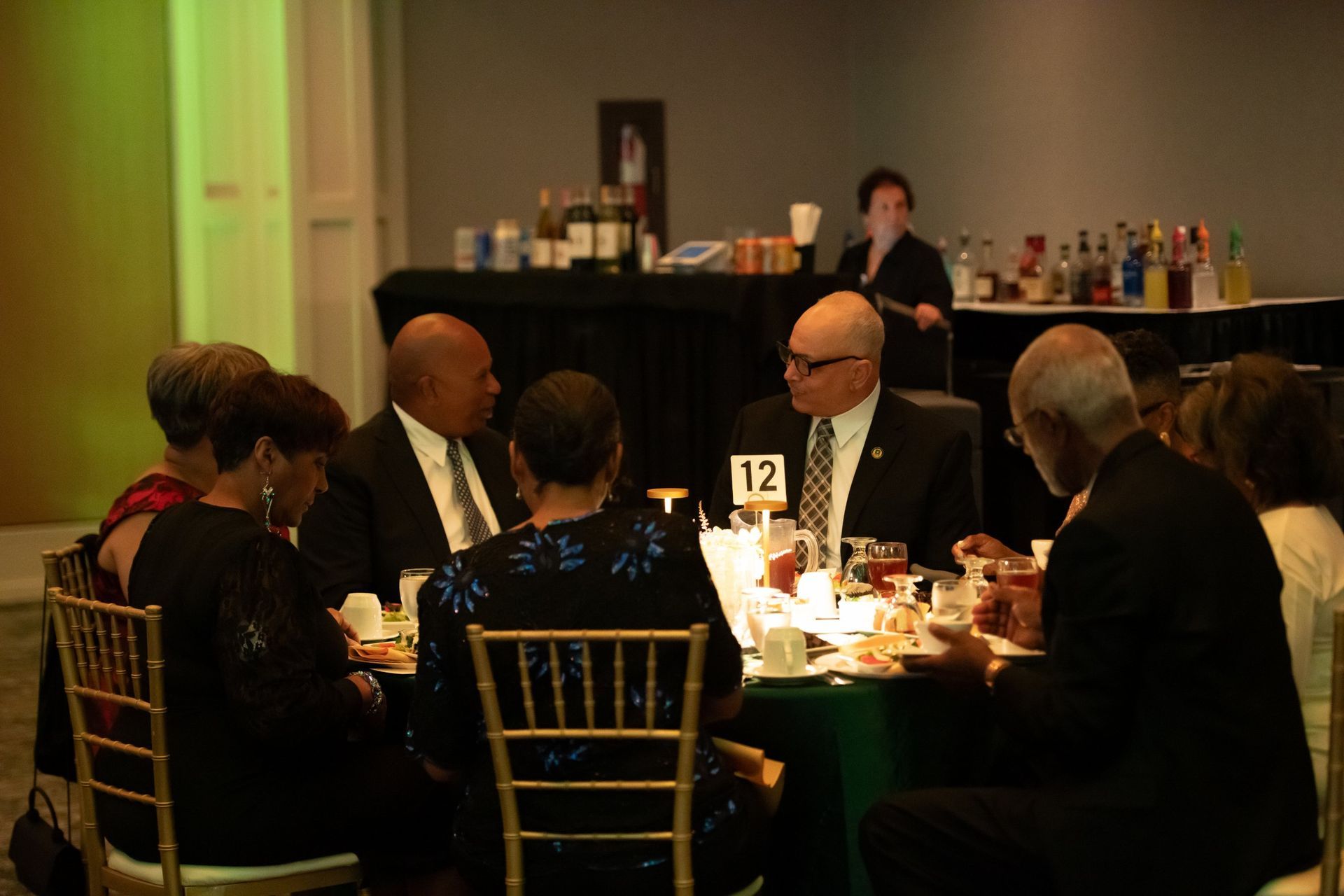 People seated at a formal banquet table listening to a speaker on a stage in the background