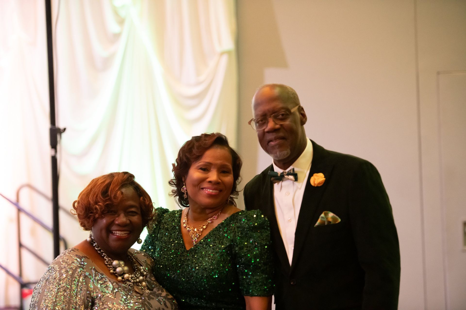 Three people in formal attire posing indoors against a white draped backdrop.