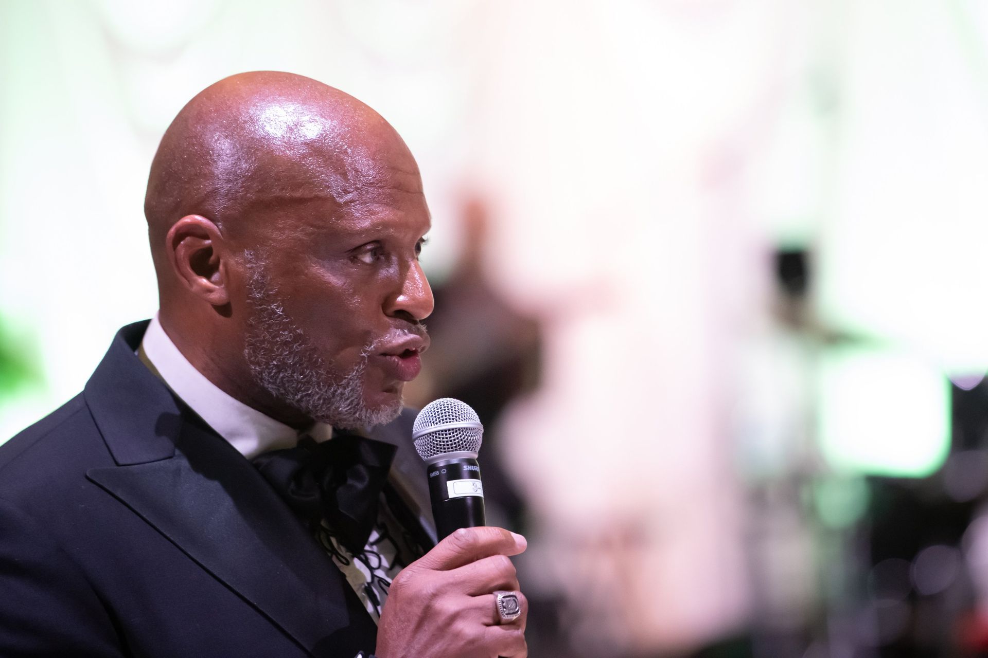 Man in a dark suit speaking into a microphone at an event, shown in profile.
