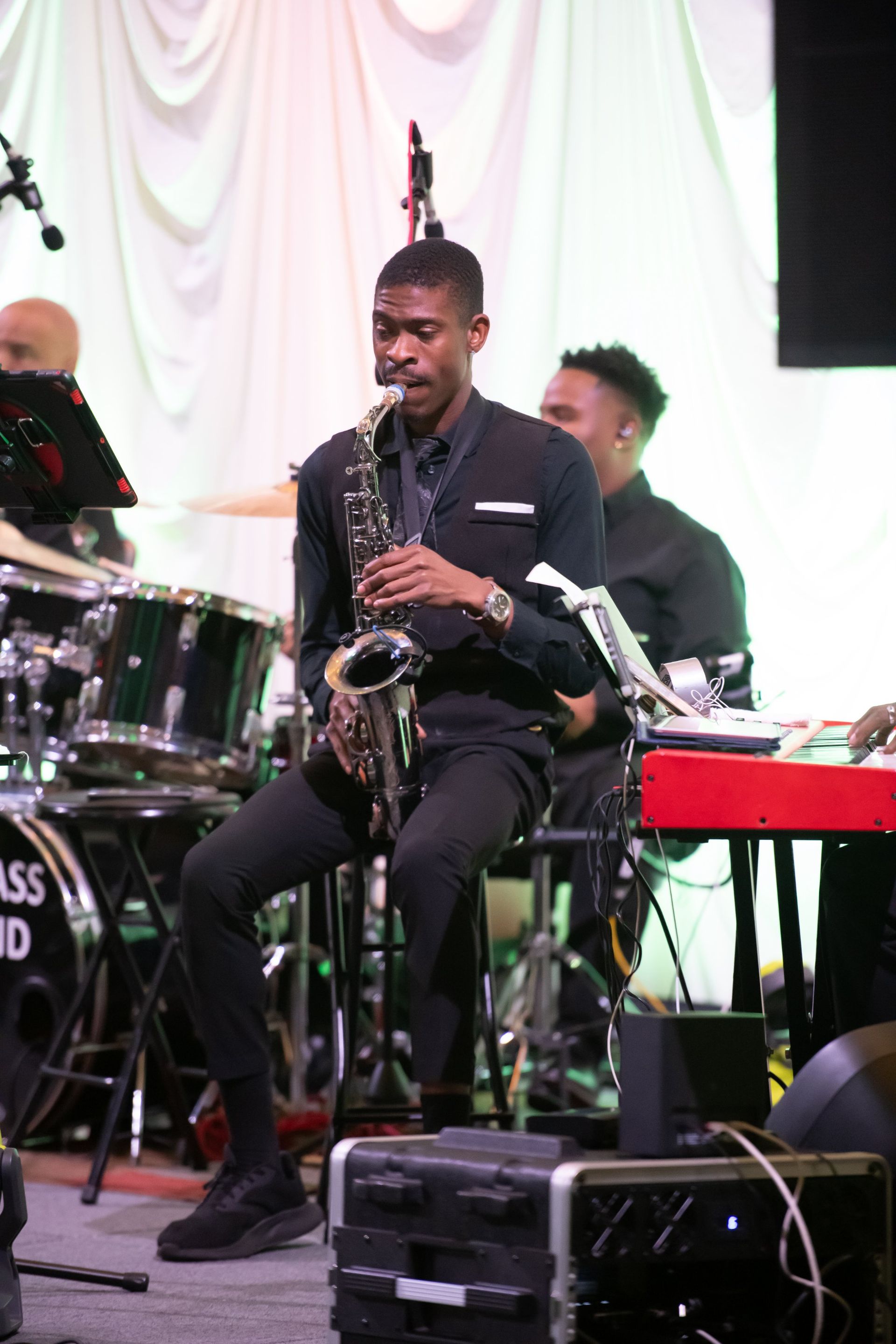 Musician playing saxophone onstage with a band, seated in front of drums and keyboard.
