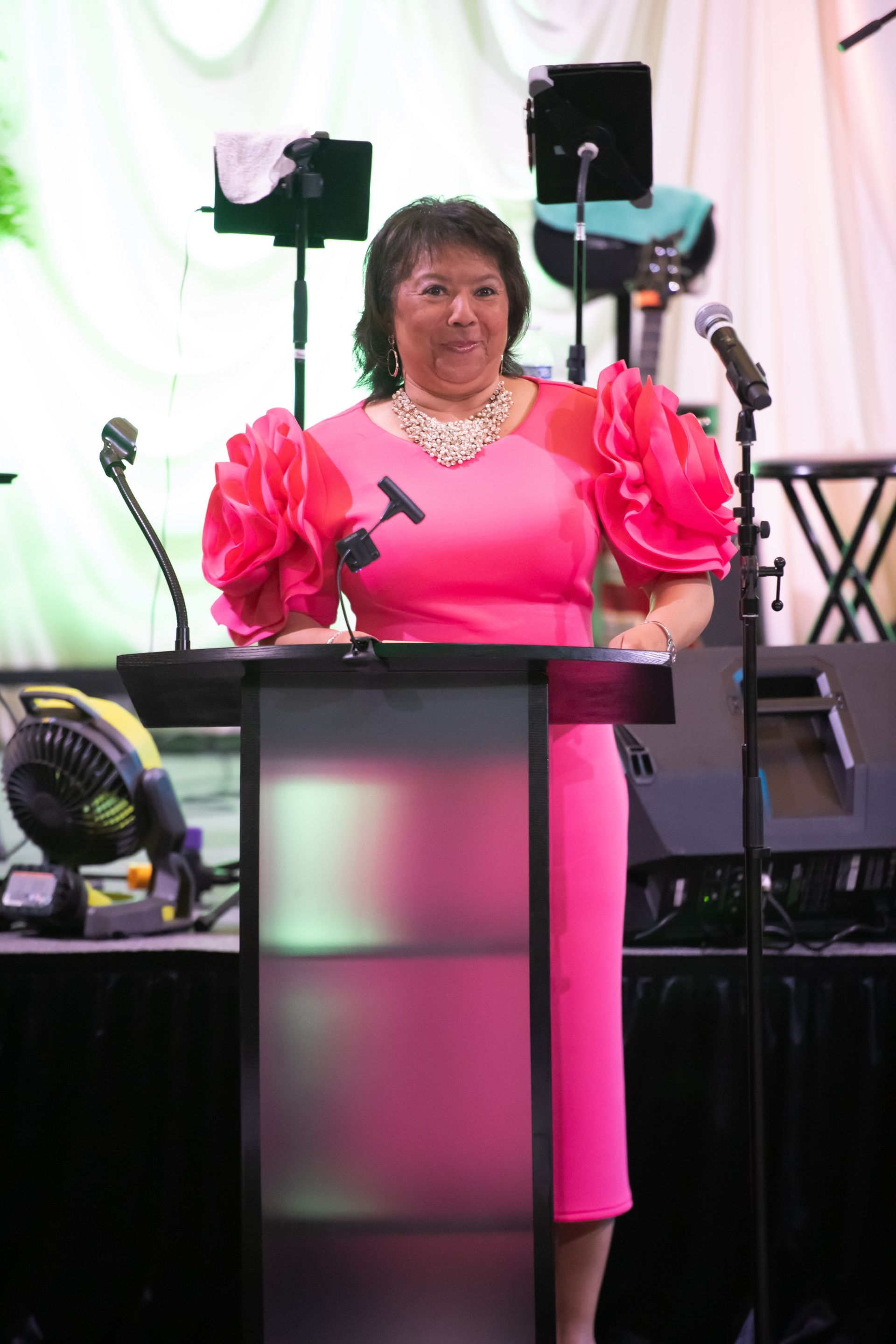 Woman in bright pink dress speaking at a podium on a stage with microphones and speakers.