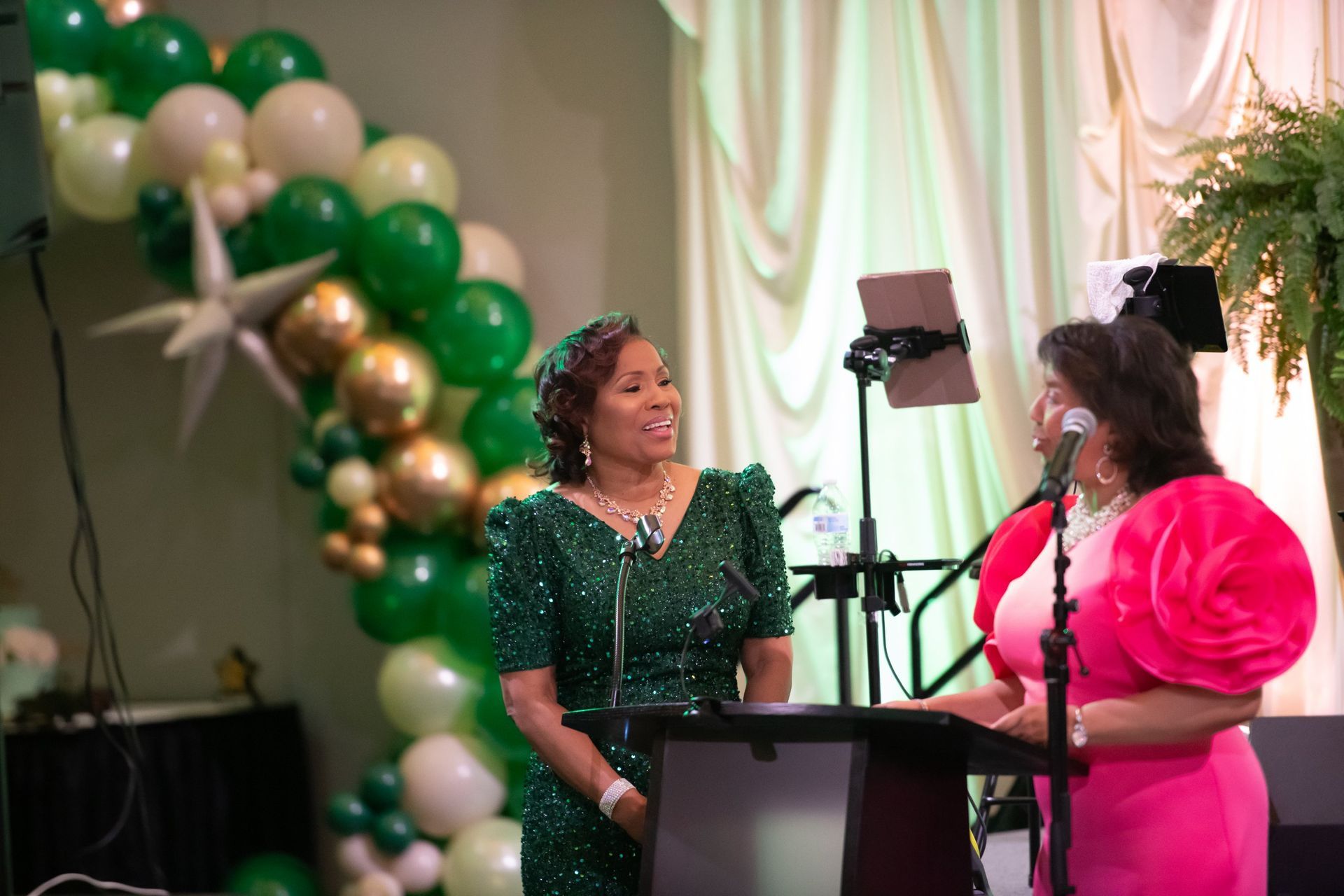 Two women at a podium in green and pink dresses, speaking at a decorated event with balloons and lights.
