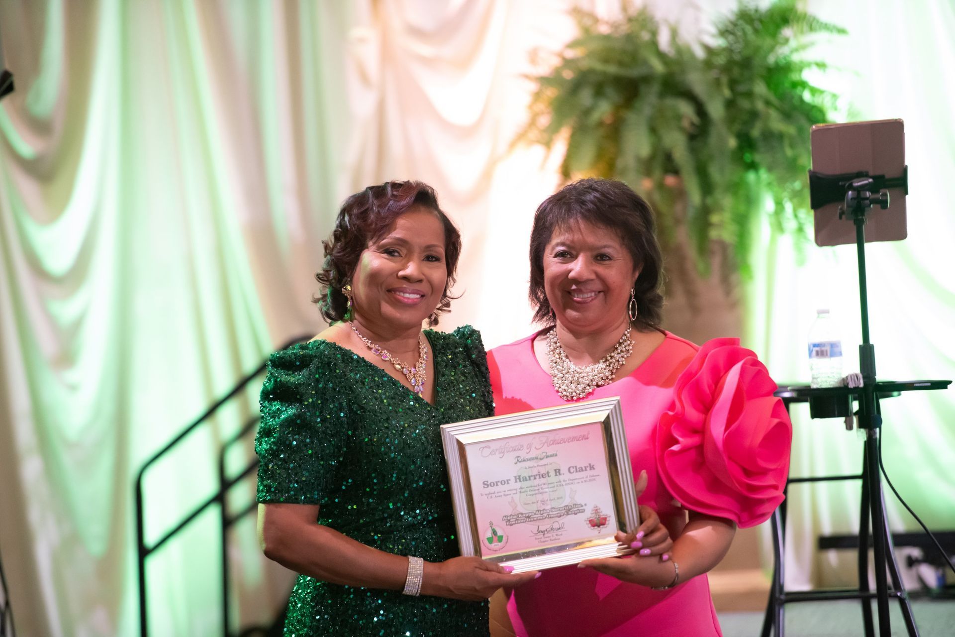 Two women in green and pink dresses hold a framed certificate at an indoor event.