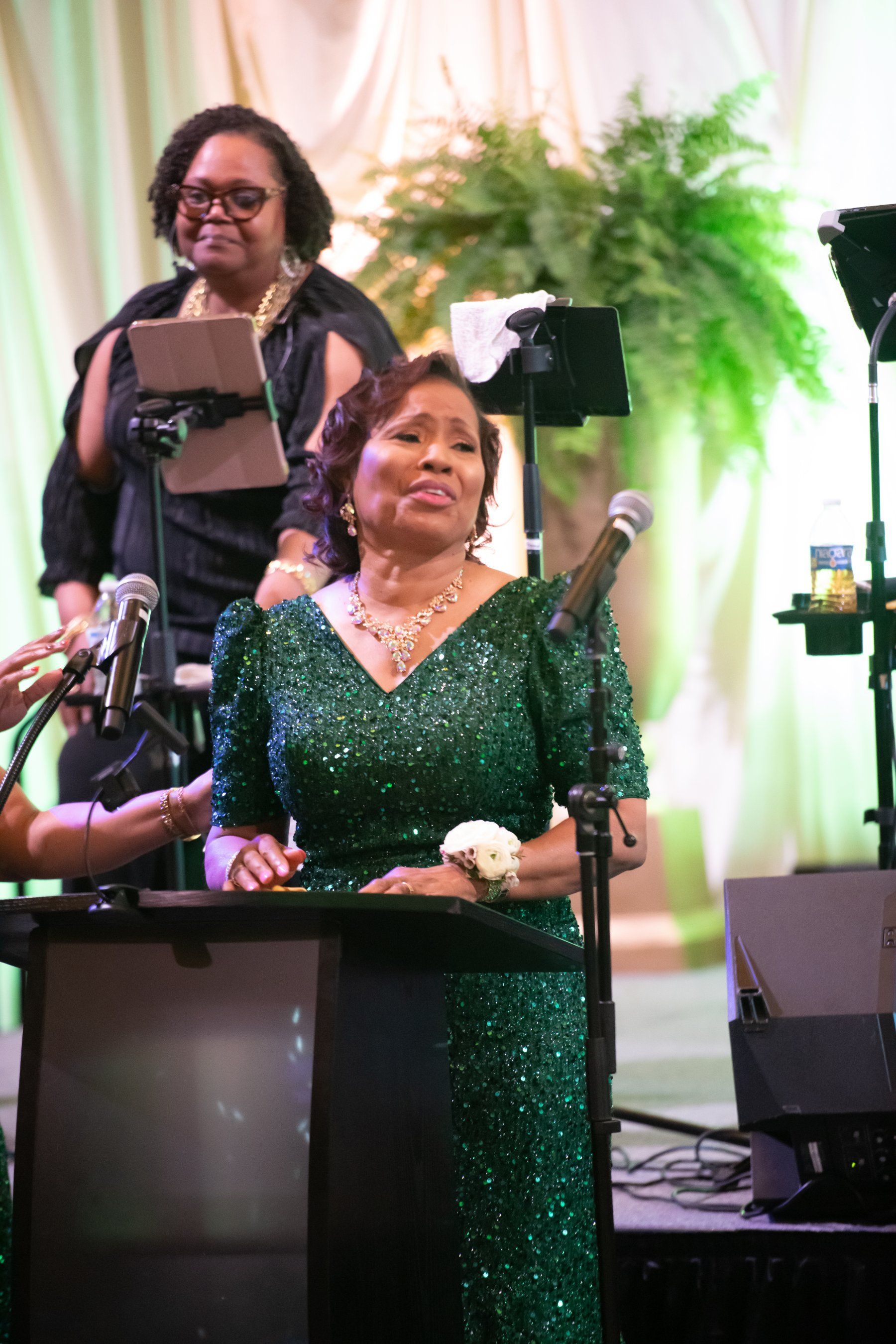 Woman in green sequined dress speaking at a podium on a stage with musicians behind her
