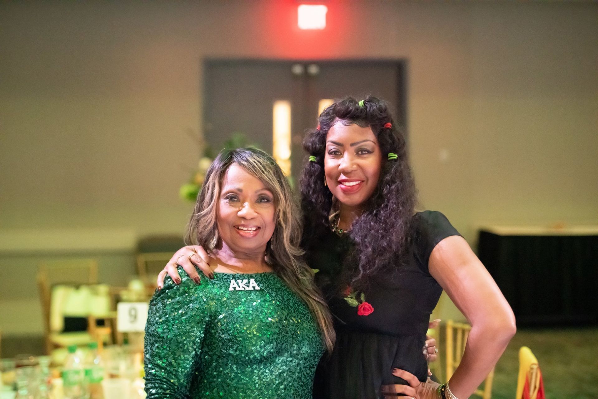 Two women posing together indoors, one in a green sequined dress and one in a black dress, both smiling.