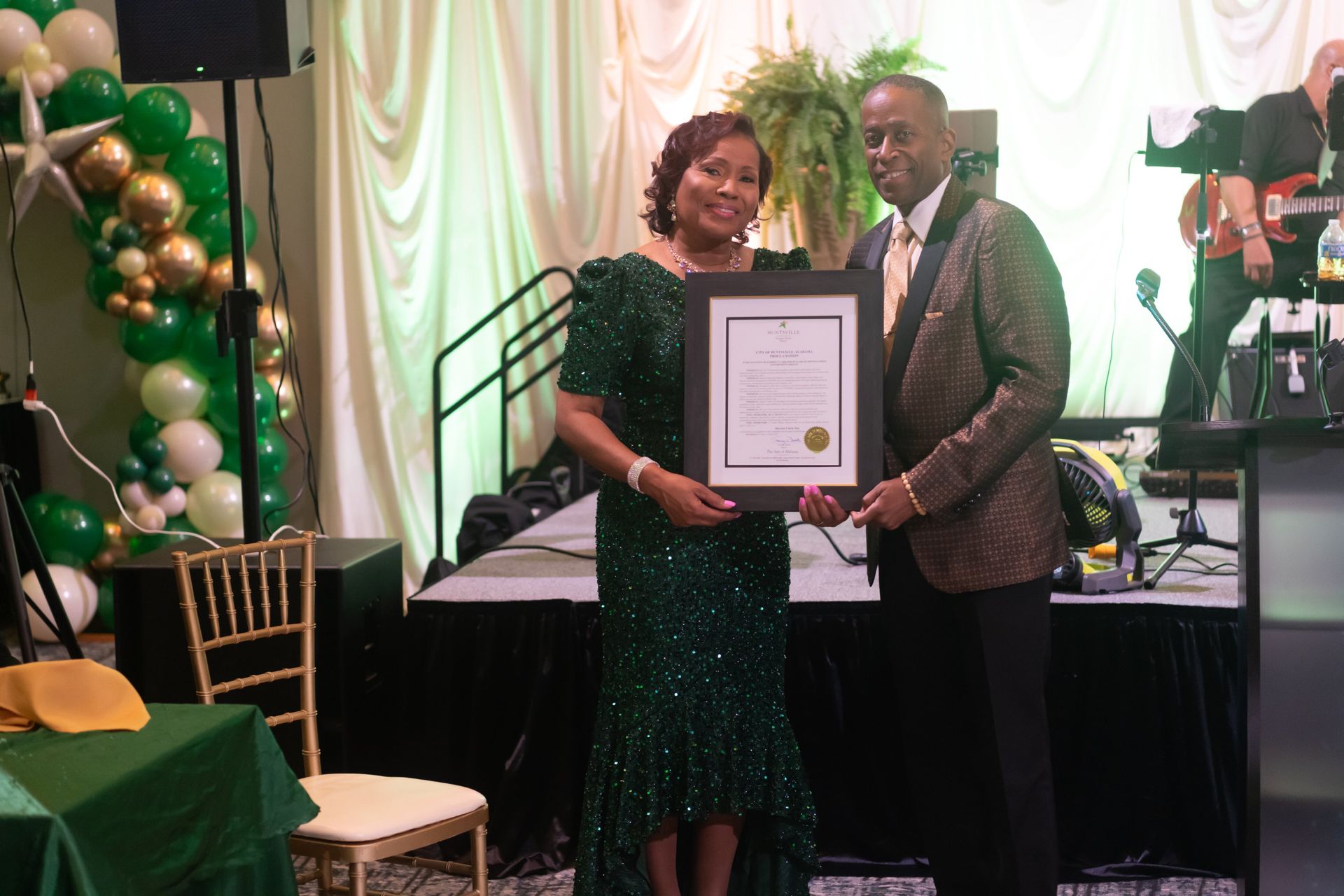 Two people holding a framed certificate at a formal awards event, with green and gold balloons behind them