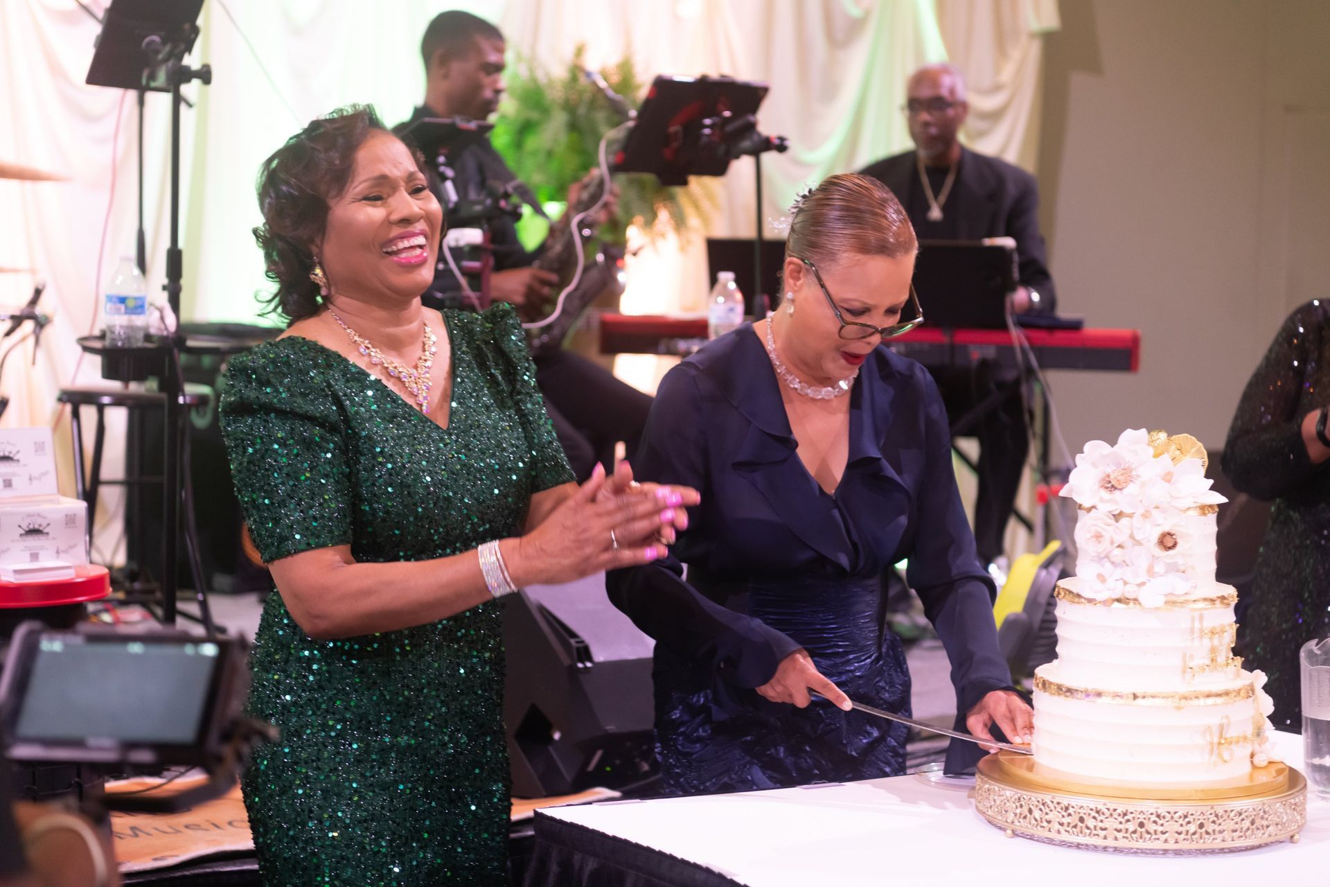 People cutting a white tiered cake at a festive event with musicians in the background