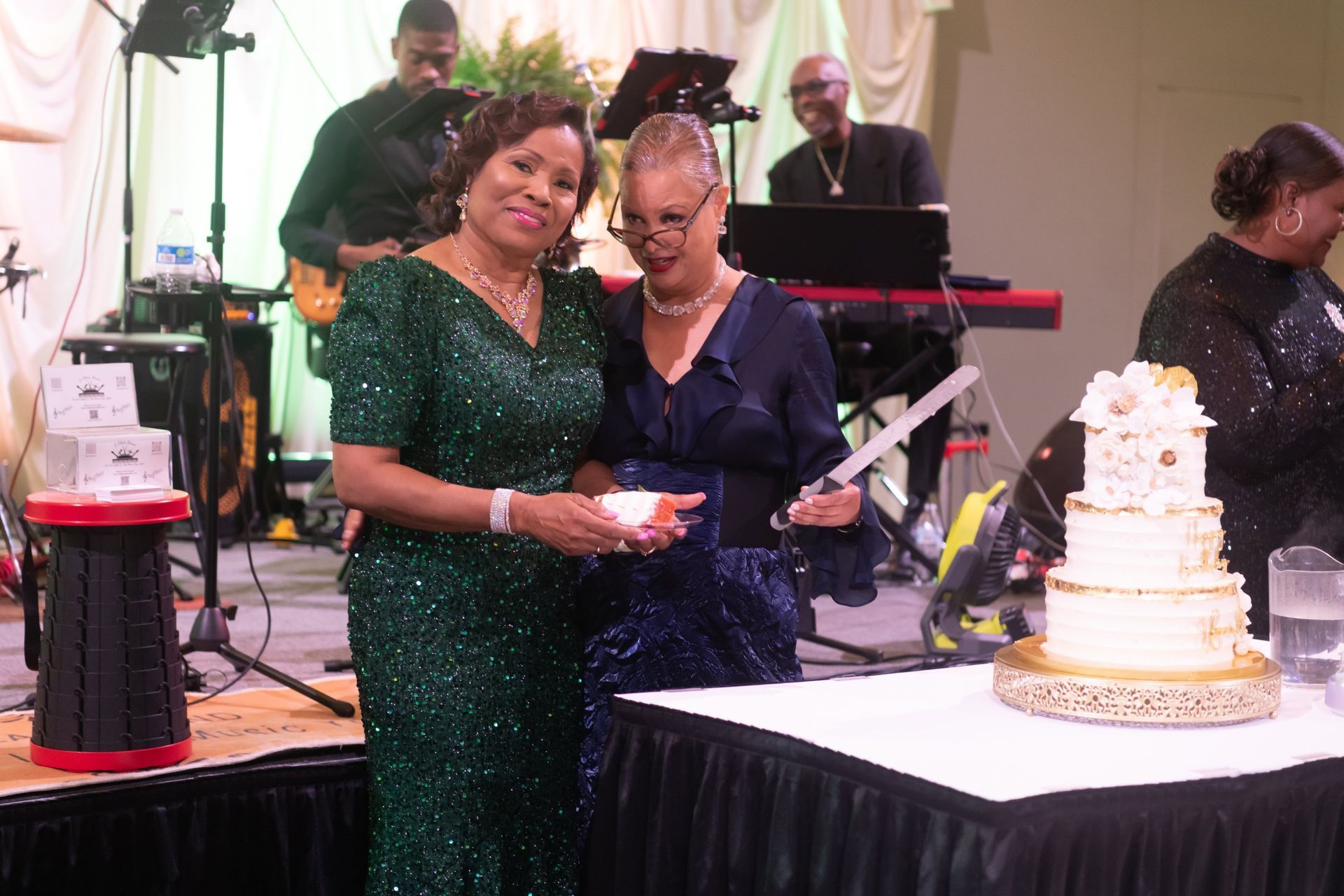 Two women at a party stand beside a white tiered cake and a table with a red drink dispenser.