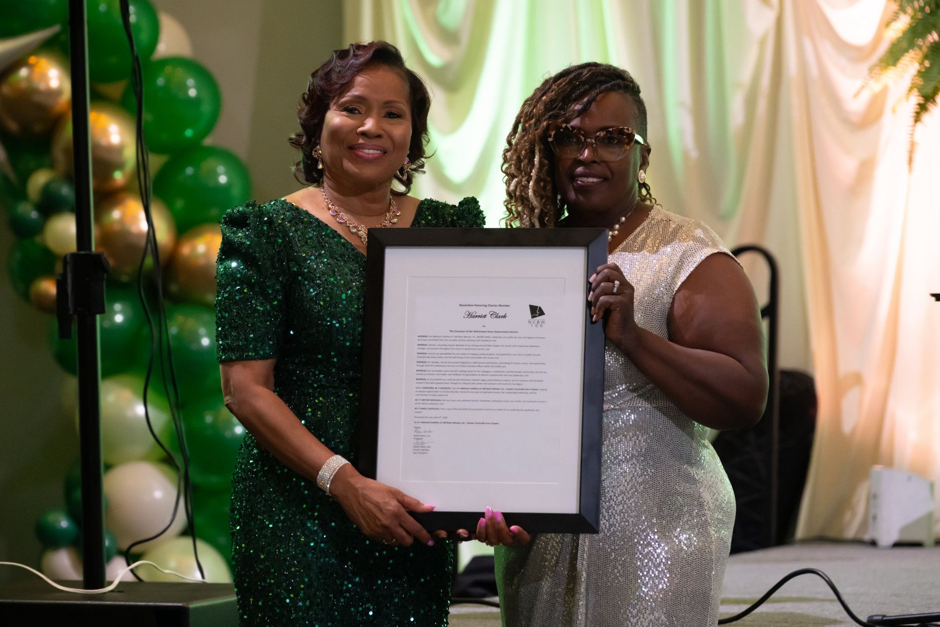 Two women in formal dresses hold a framed certificate at a green-themed ceremony.