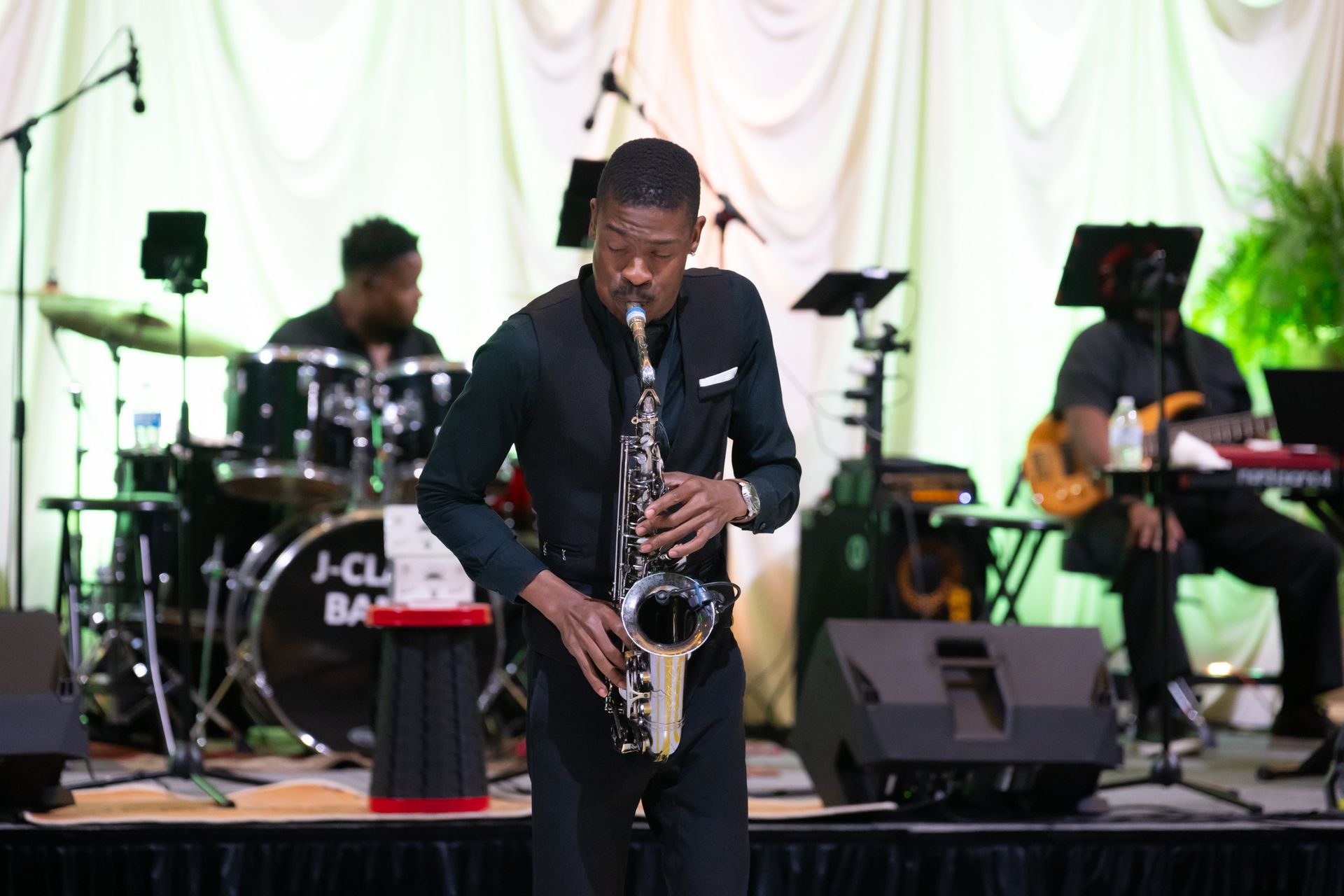 Saxophonist performing onstage with a live band, in front of white draped curtains and speakers.