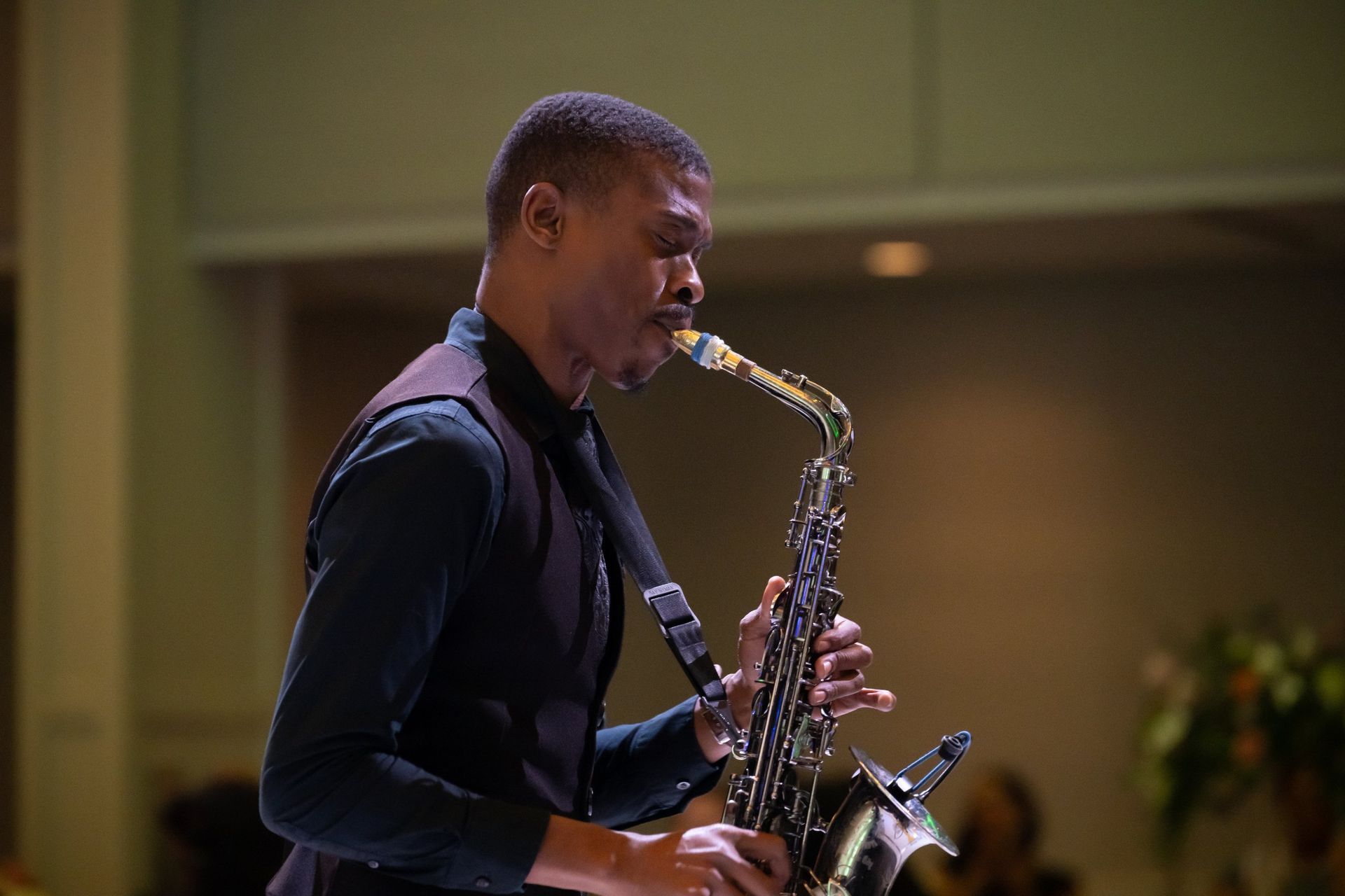 Musician playing saxophone on stage in a dark suit, lit by warm indoor lighting.