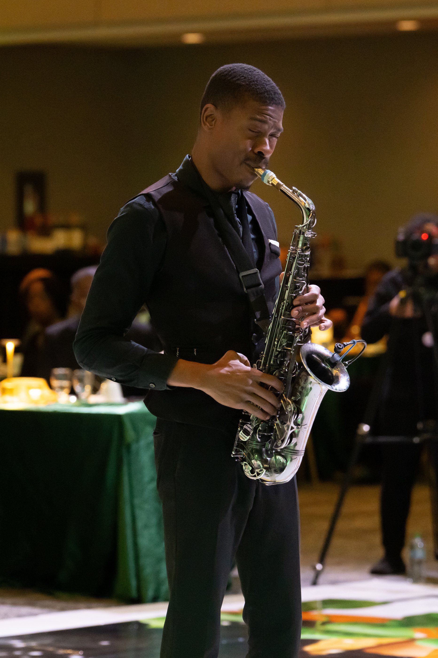 Man playing a saxophone at an indoor event, wearing a dark shirt and black pants